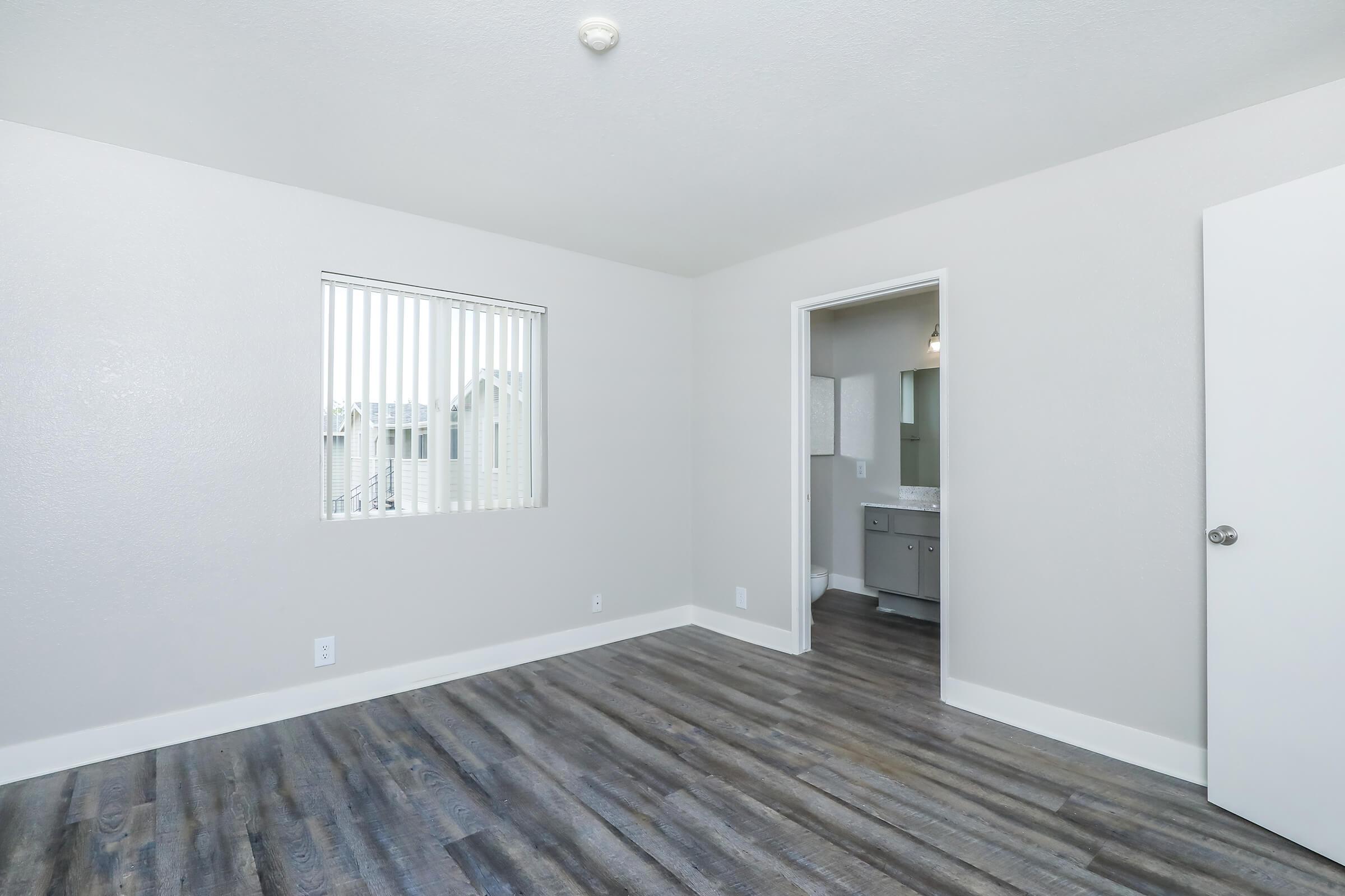 A spacious, empty room with light grey walls and wood-style flooring. A window with vertical blinds allows natural light, and a doorway leads to an adjacent bathroom. The room has a neutral and modern feel, ideal for decorating or furnishing.