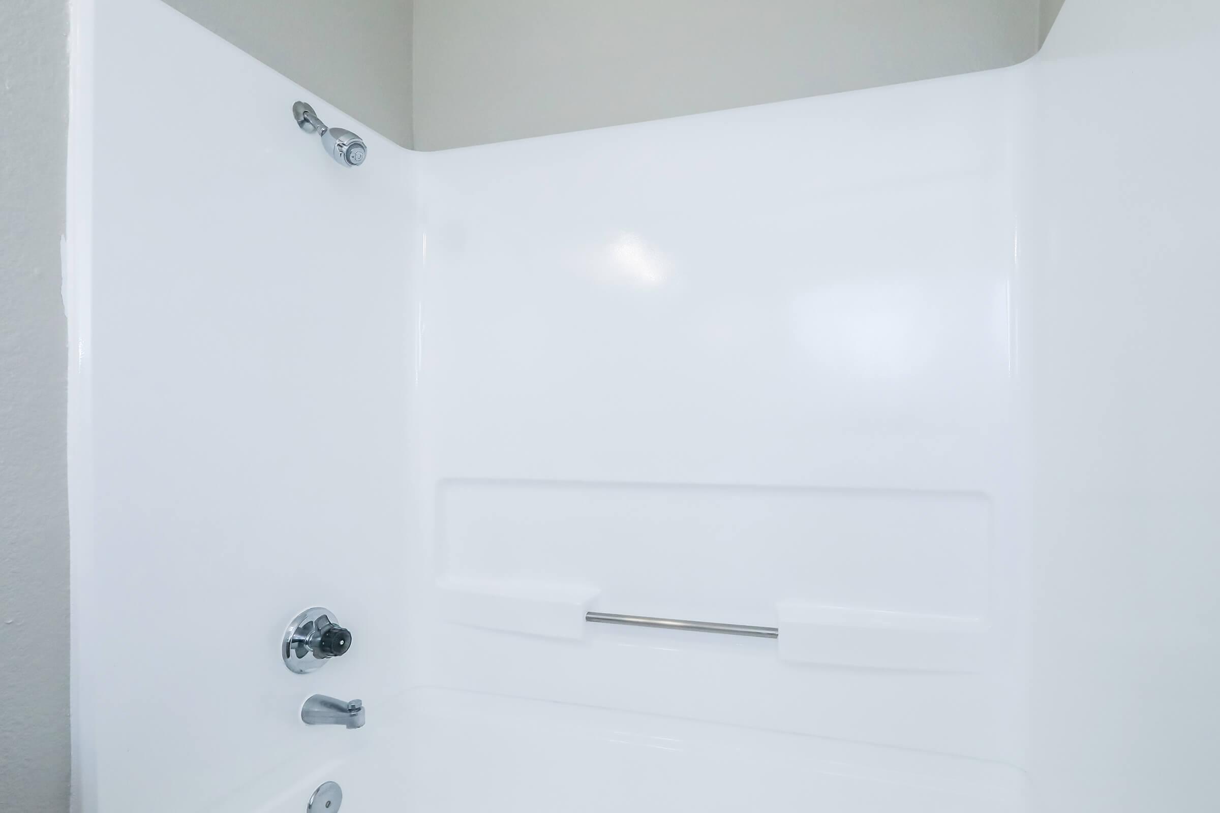 A clean, modern shower enclosure with white, smooth walls and a chrome faucet and showerhead. The space features a grab bar and is well-lit, creating a fresh and inviting atmosphere.