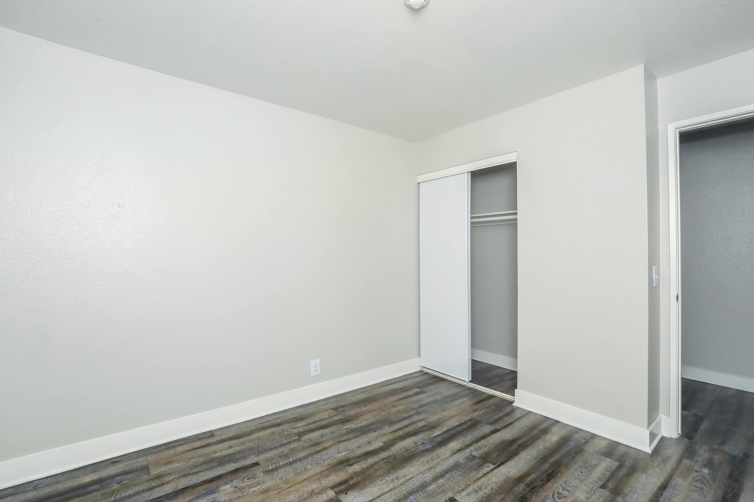 Empty room with light-colored walls and a wooden floor. A sliding closet door is visible on the left side, and there's a doorway on the right leading to another space. The overall atmosphere is bright and minimalistic, suitable for a bedroom or office.