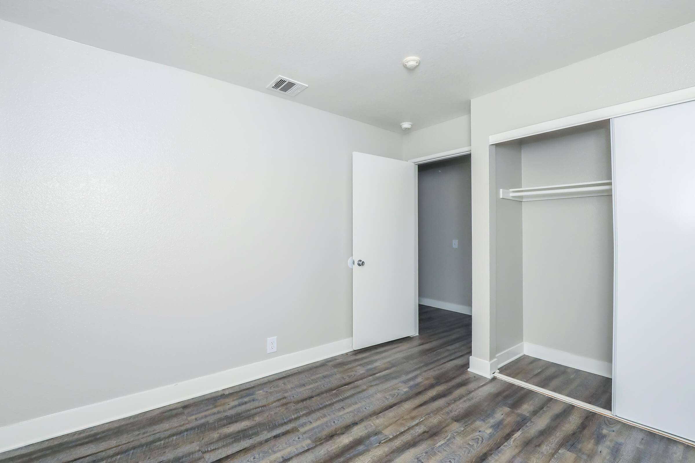 A vacant bedroom featuring light gray walls and wood laminate flooring. The space includes a closed door leading to another room and a closet with a sliding door that is partly open, showcasing an empty interior. The overall atmosphere is clean and minimalist.