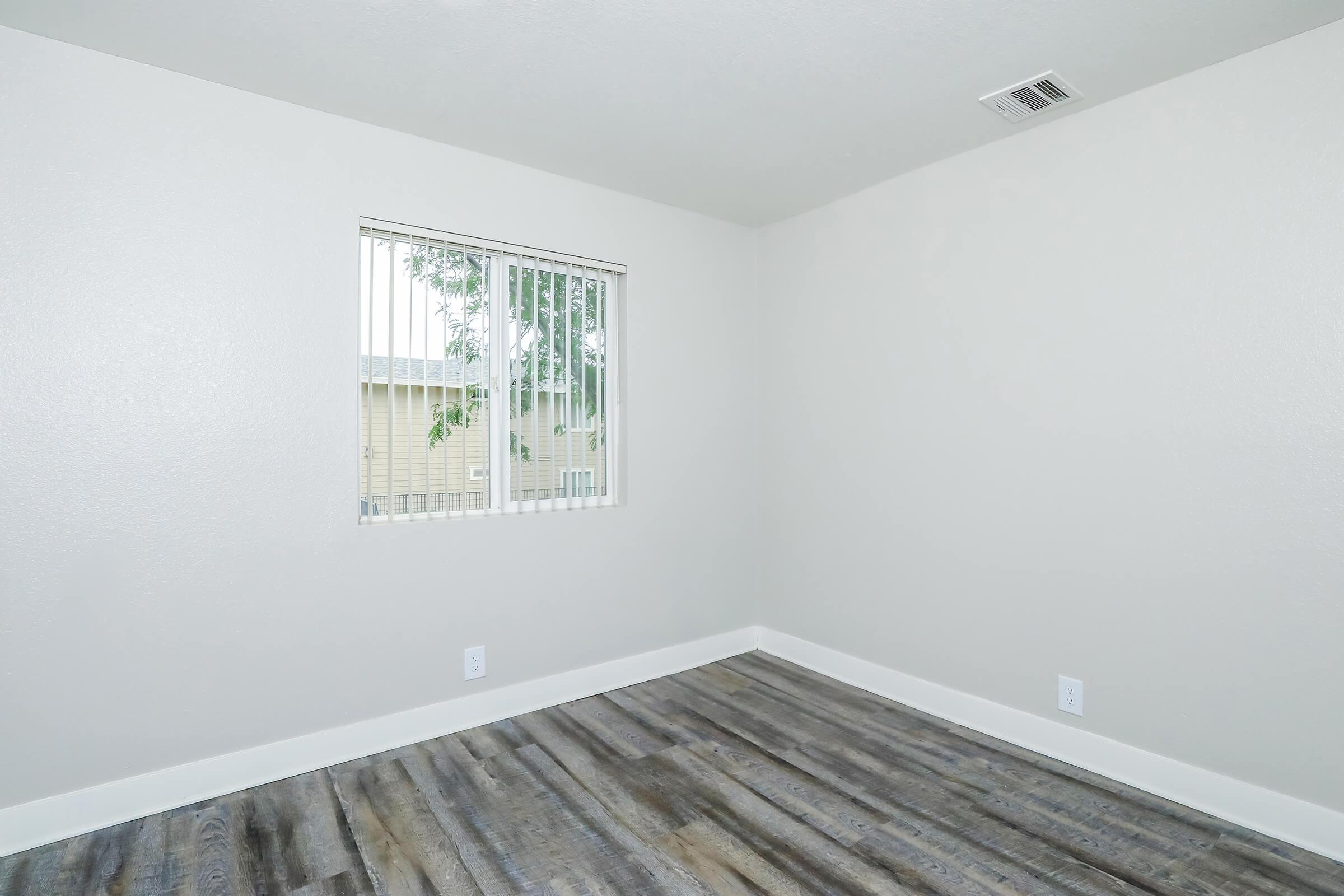 Empty room featuring light gray walls and wood-like flooring. A window with vertical blinds allows natural light to enter. The space is unfurnished, providing a blank canvas for decoration or use.