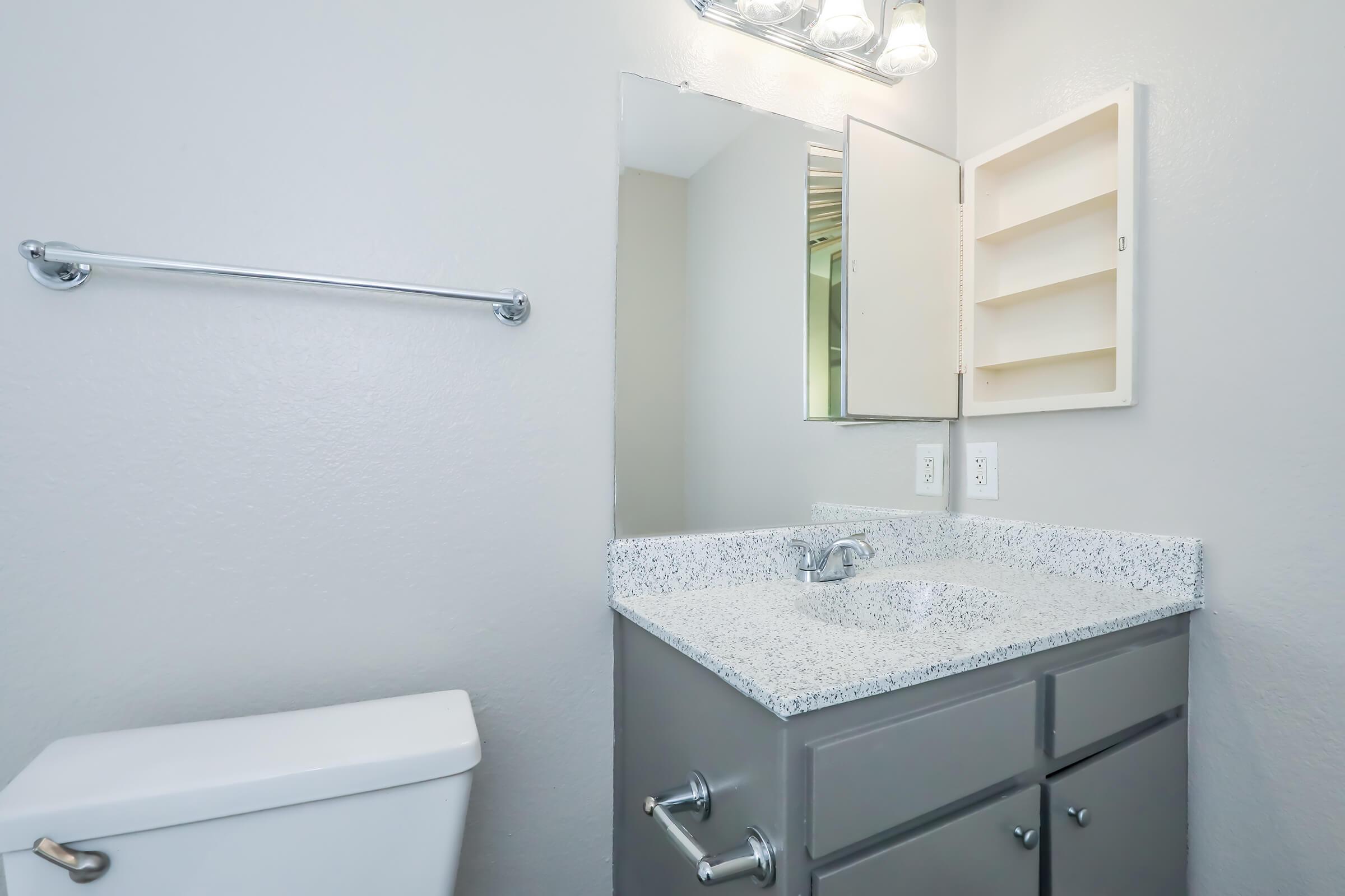 A modern bathroom featuring a gray vanity with a white speckled countertop, a mirror above the sink, and a small cabinet. A white toilet is visible on the left, with a towel rack on the wall. The walls are painted in a light gray color, contributing to a clean and simple aesthetic.