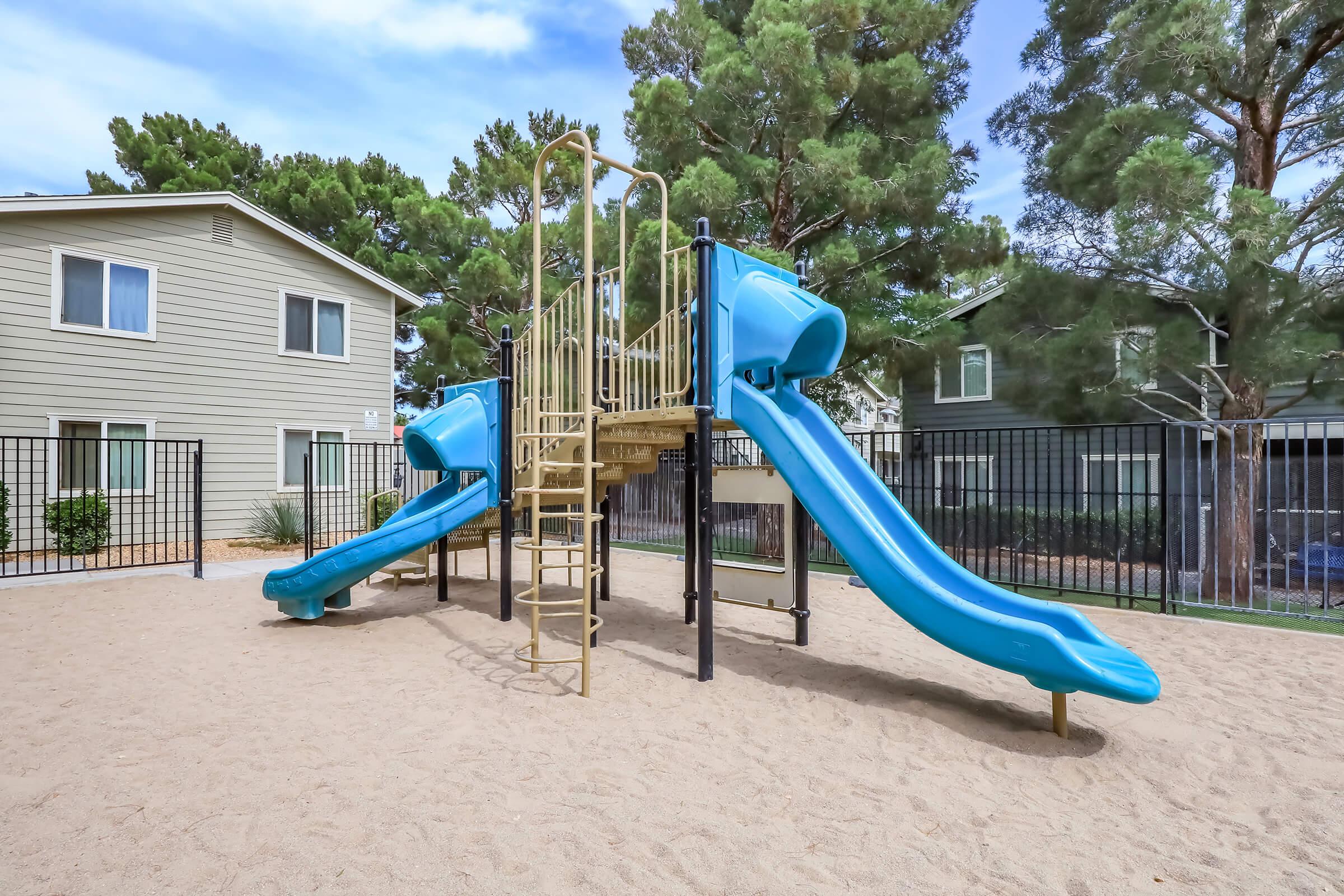 A playground featuring two blue slides and a climbing structure, surrounded by a sandy area and backed by green trees and residential buildings. The area is enclosed with a black fence, providing a safe play environment for children.