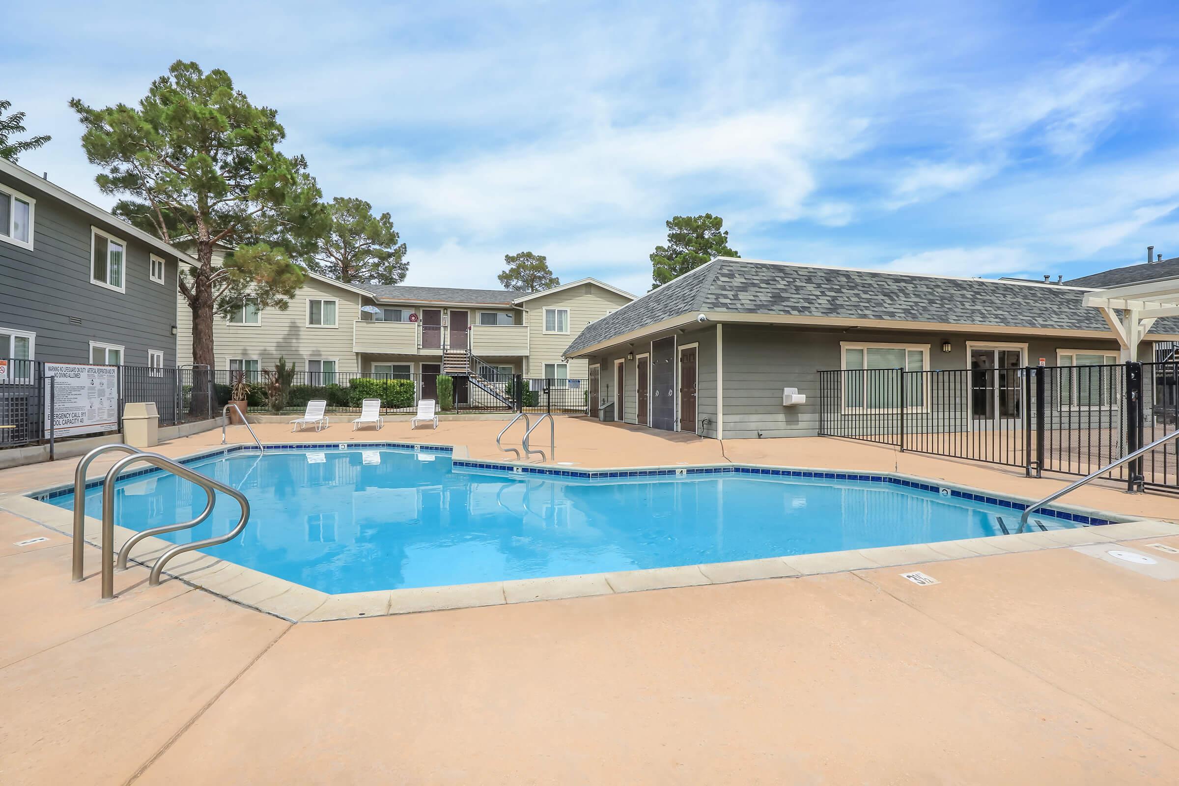 A clear blue swimming pool surrounded by a sandy deck, with white lounge chairs nearby. In the background, two multi-story residential buildings with green trees and a cloudy sky are visible. A fenced area encloses the pool, providing a tranquil outdoor space.