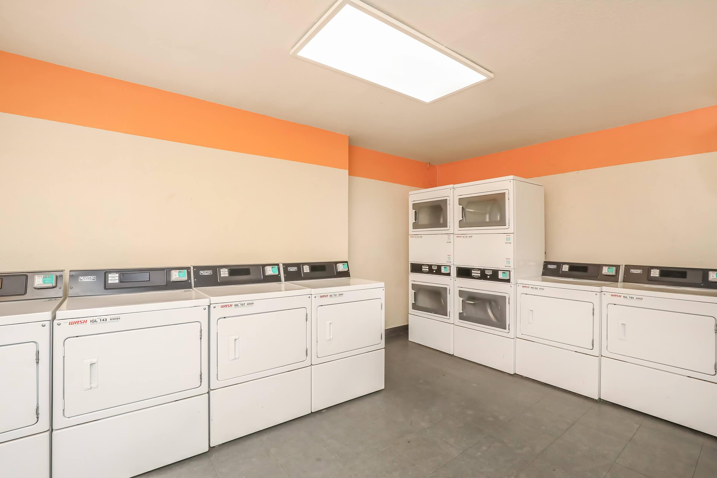 A clean and bright laundromat featuring several white washing machines and dryers arranged along the walls. The space has a light-colored floor and orange accent walls, with a ceiling light providing ample illumination. There are no people present in the image.