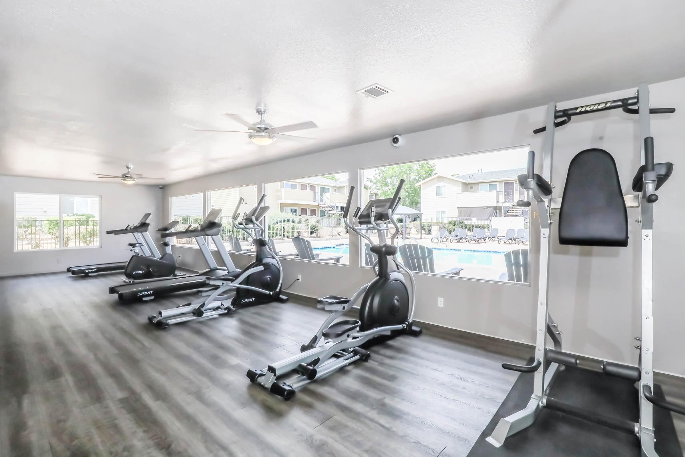 A bright and spacious fitness room featuring several modern exercise machines like ellipticals and stationary bikes. Large windows provide natural light and a view of a pool area outside. The room has wooden flooring and ceiling fans for ventilation.