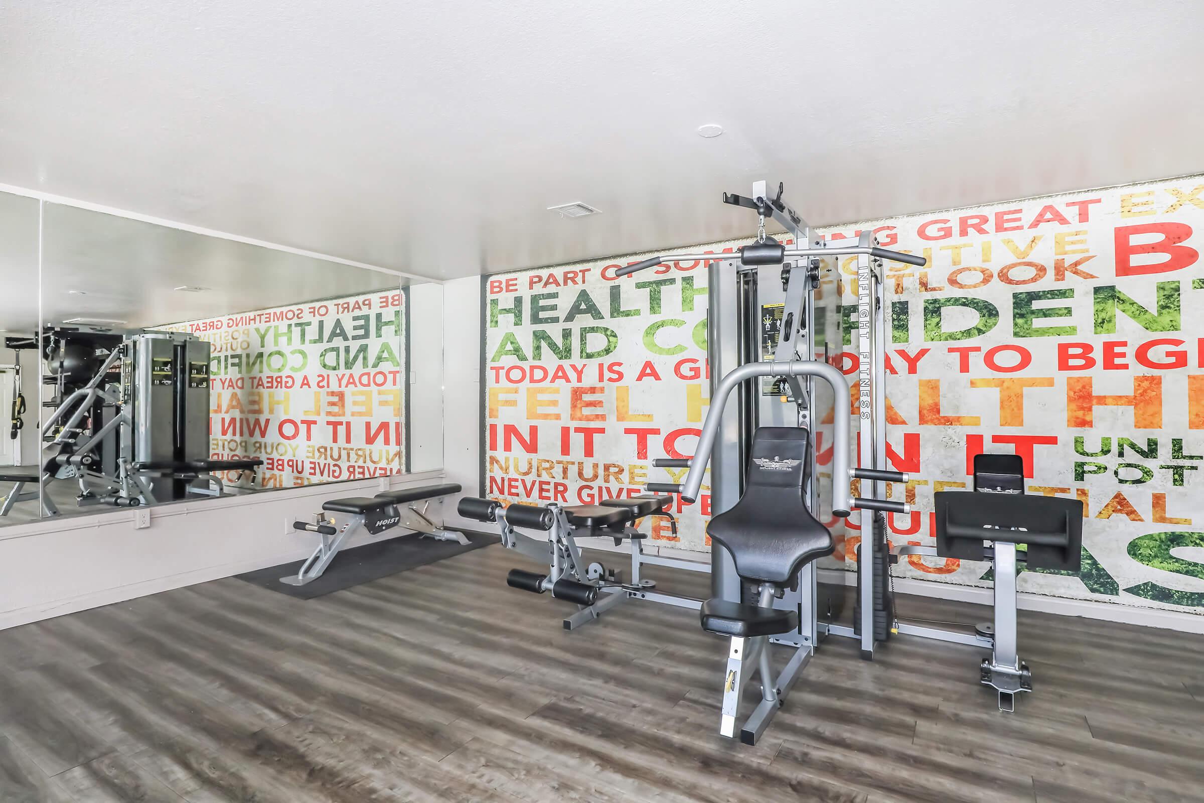 A modern gym interior featuring exercise equipment, including a weight machine and bench, set against a colorful motivational wall with phrases about health and wellness. A large mirror reflects the space, creating an open and inviting atmosphere for workouts.