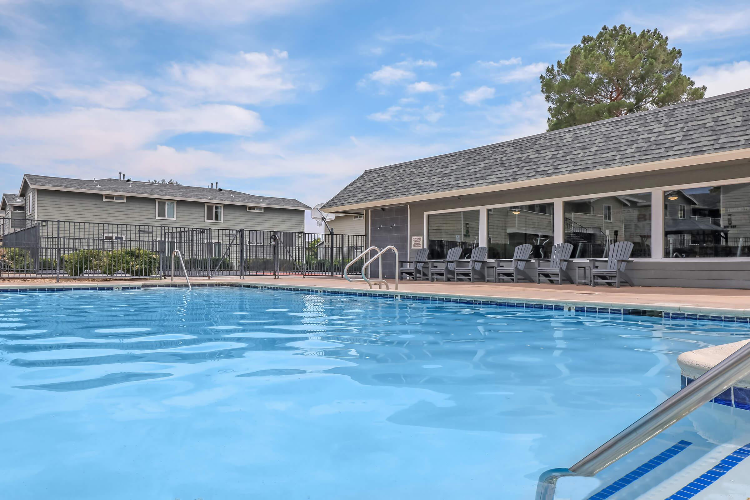 A clear blue swimming pool with steps leading in, surrounded by a wooden deck. In the background, a building with large windows and a shingled roof is visible, along with several black lounge chairs. The scene is set against a cloudy sky and green trees.