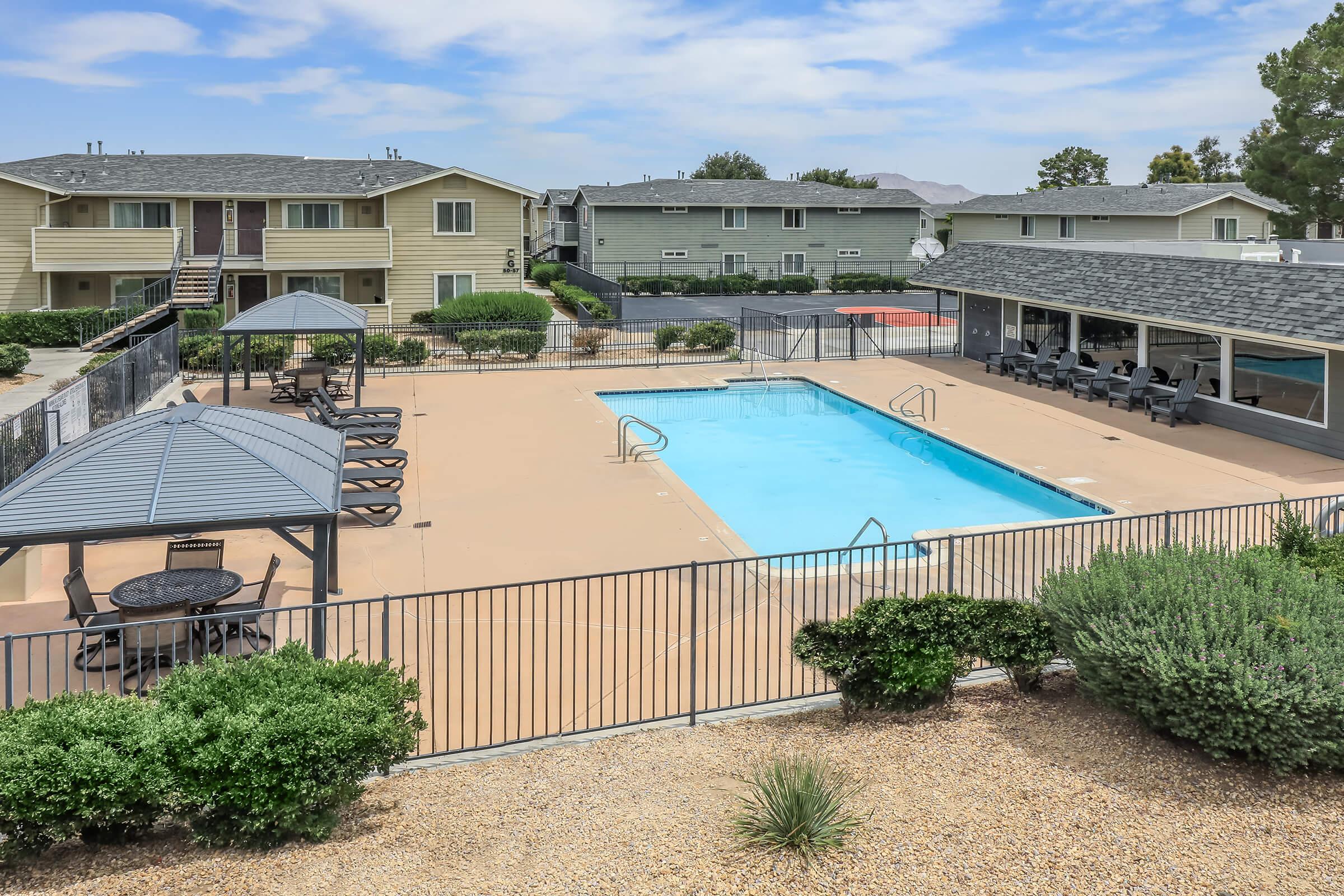 A spacious outdoor area featuring a swimming pool surrounded by a sun deck with lounge chairs. There are shaded seating areas with tables and a basketball court visible in the background. The setting includes well-maintained landscaping and residential buildings nearby, under a clear blue sky.
