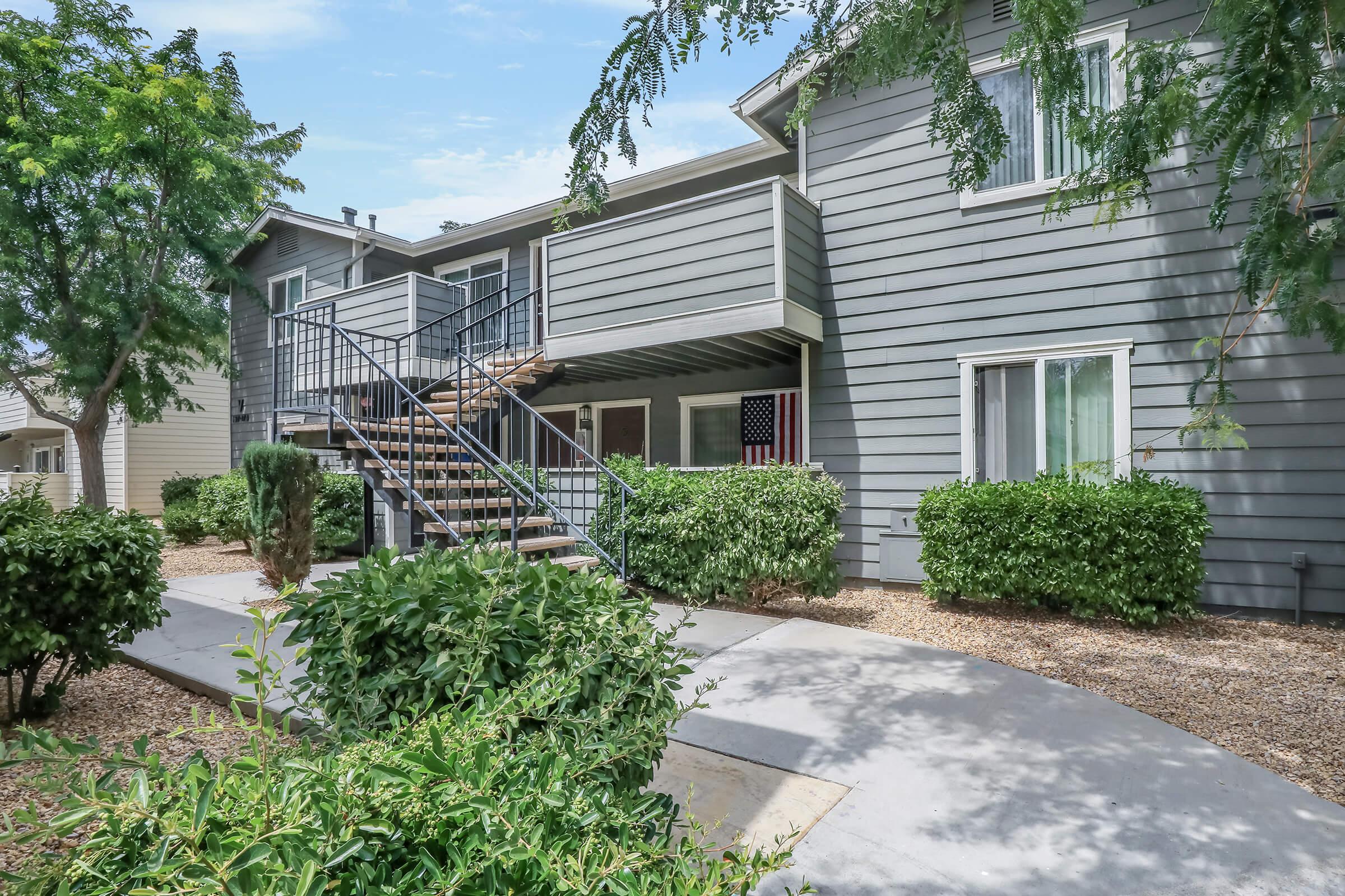 Two-story residential building with gray siding, featuring a staircase leading to an upper level. The pathway and surrounding area are landscaped with shrubs and small trees. Clear blue sky overhead, creating a bright and inviting atmosphere.