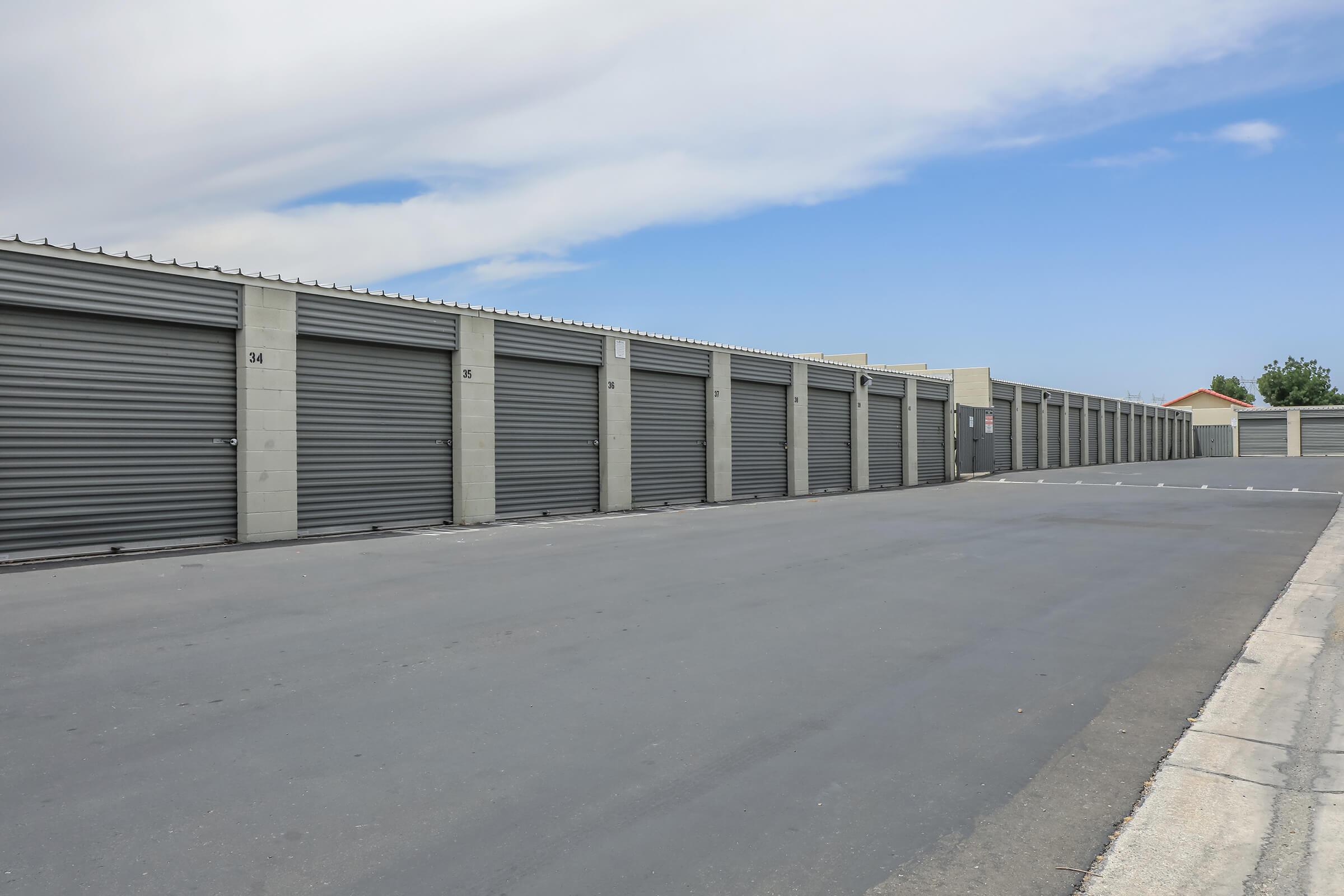 A row of storage units with metal gray doors, set against a clear blue sky. The surrounding area features a clean asphalt surface and a few scattered plants. The scene conveys a sense of organization and accessibility in a storage facility.