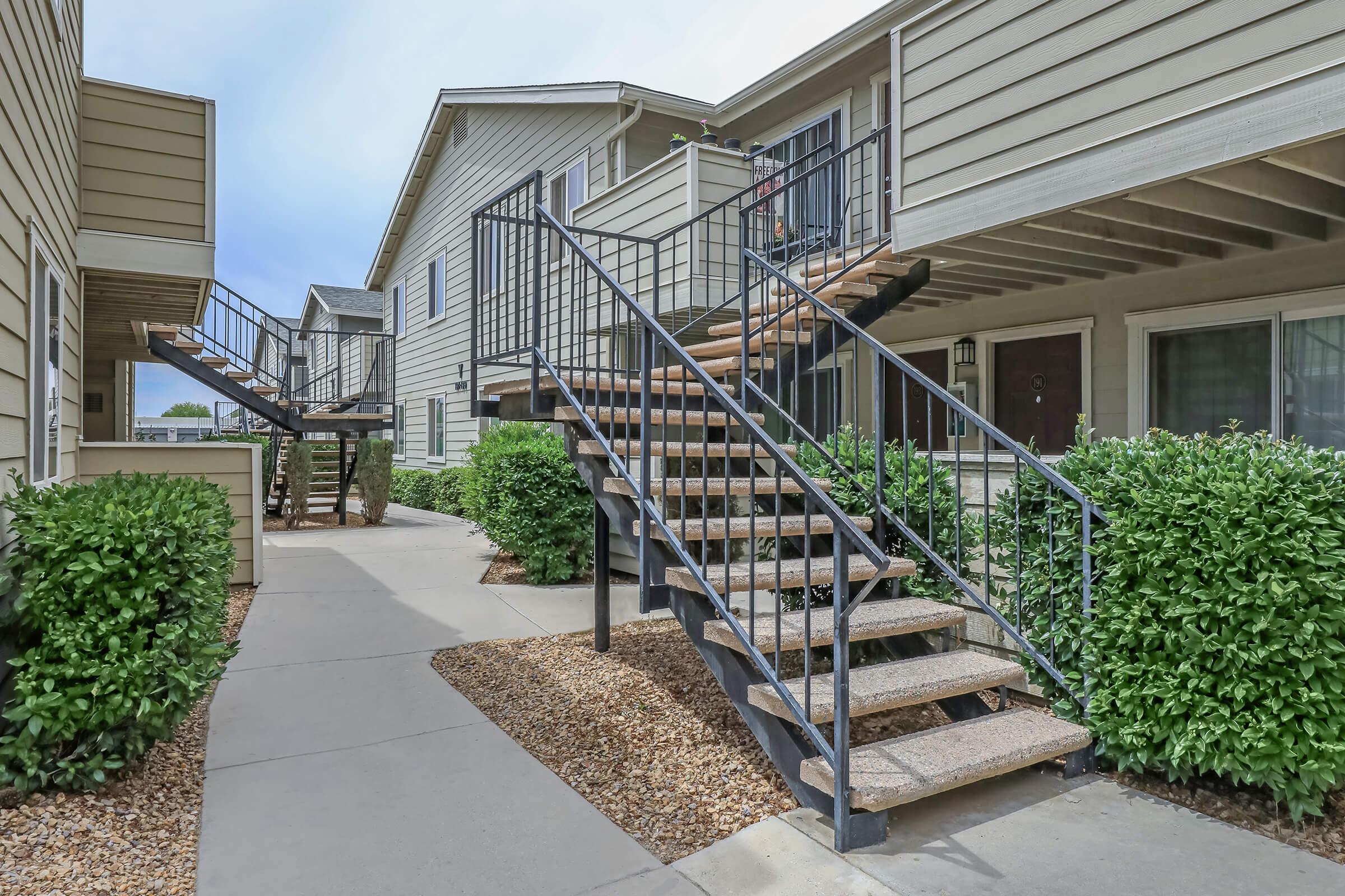 A view of an outdoor apartment complex pathway featuring two sets of metal stairs leading up to upper-level units. The pathway is lined with neatly trimmed hedges and gravel landscaping, with two adjacent buildings visible in the background under a partly cloudy sky.