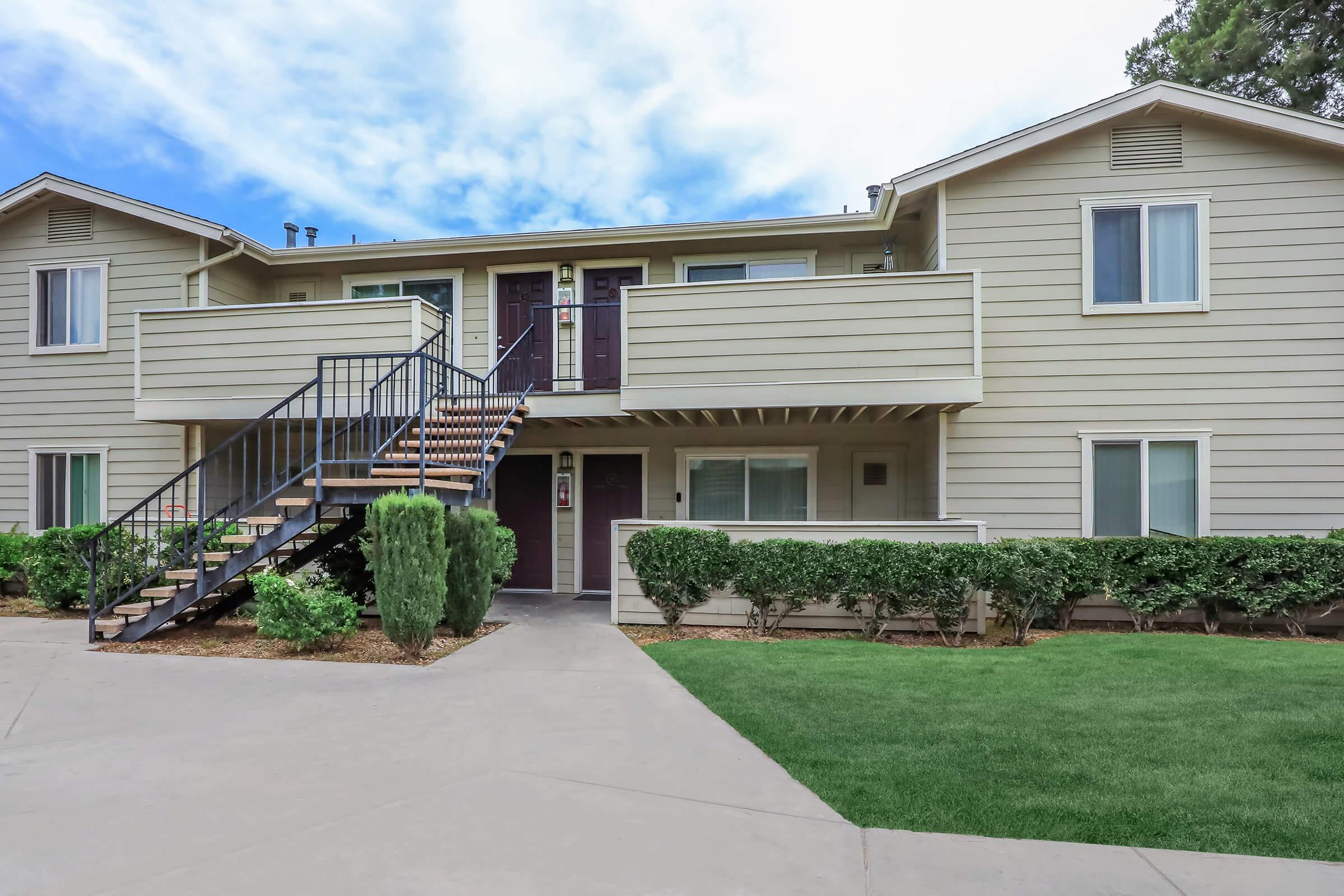 A two-story apartment building with beige siding, featuring balconies on each unit. The entrance has a set of stairs leading up to the second floor. There are well-maintained shrubs and grass in the foreground, and a bright blue sky with clouds in the background.