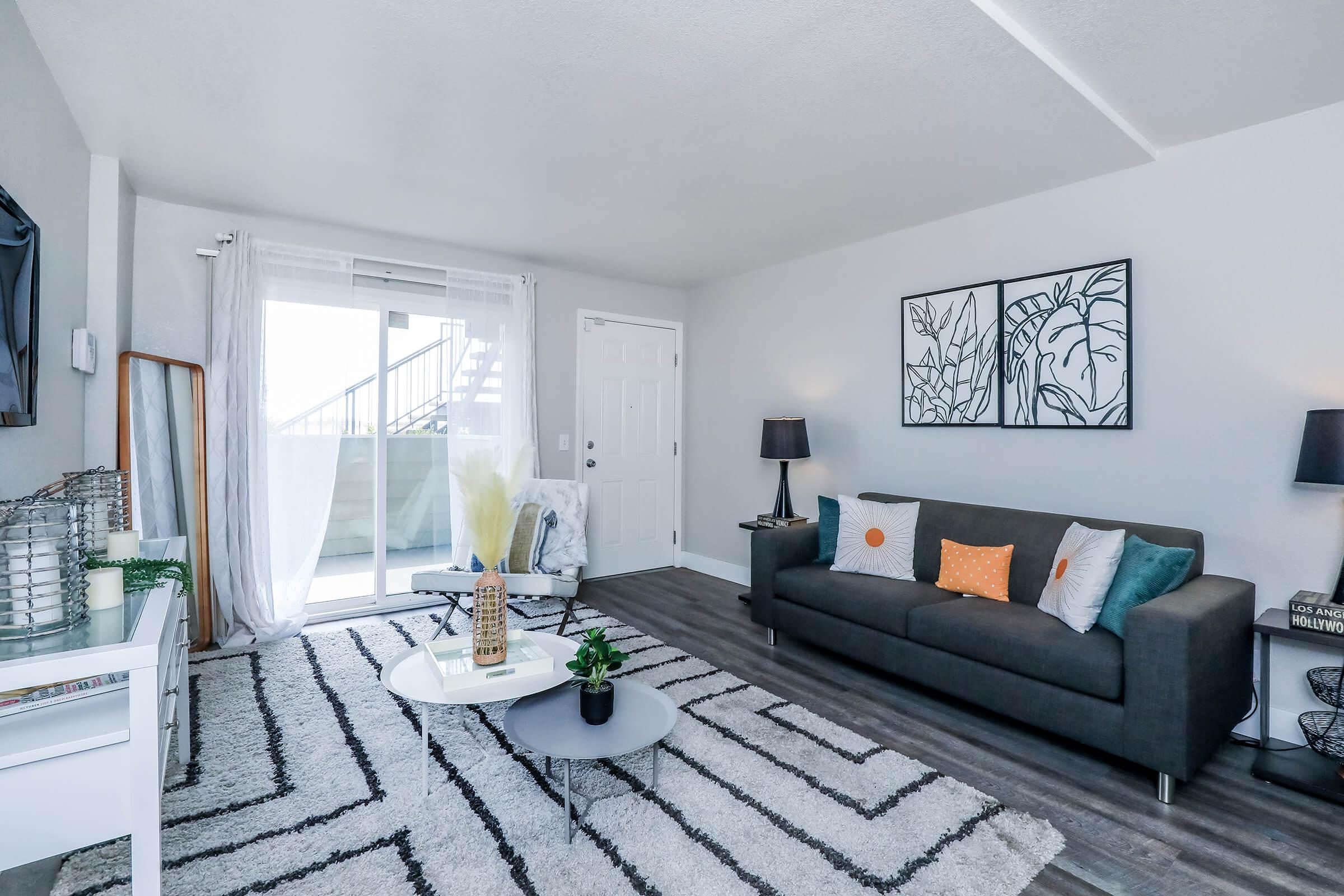 A modern living room featuring a gray sofa with colorful throw pillows, a round coffee table, and a patterned area rug. There are potted plants, a TV mounted on the wall, and large windows with sheer curtains allowing natural light. Wall art with abstract designs adds decoration to the space.