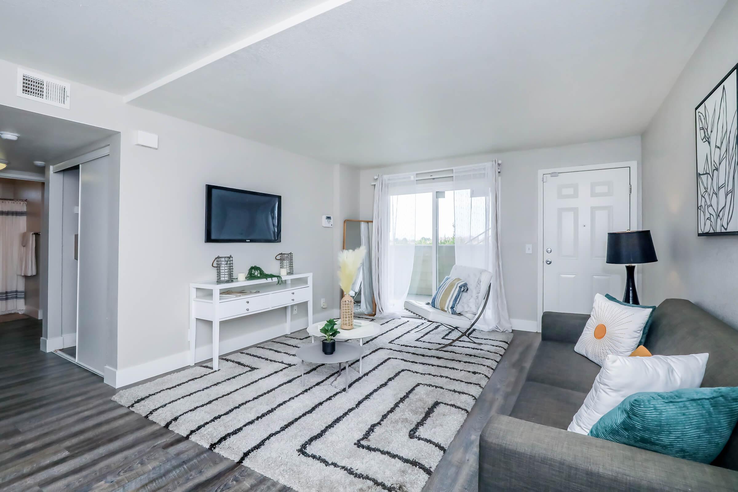 A modern living room featuring a gray sofa with decorative pillows, a glass coffee table on a striped rug, and a flat-screen TV on the wall. Large windows provide natural light, and there is a balcony door. Decorative plants and a lamp complete the cozy atmosphere.