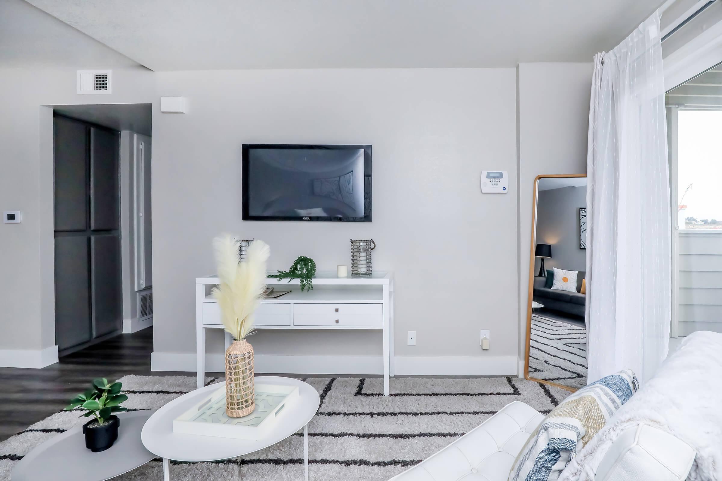 A modern living room featuring a white console table with a decorative vase and plant, a wall-mounted TV, a large mirror, and a plush sofa. The walls are painted in soft gray, and there is a light area rug on the floor, creating a cozy and inviting atmosphere. Natural light streams in from a window.