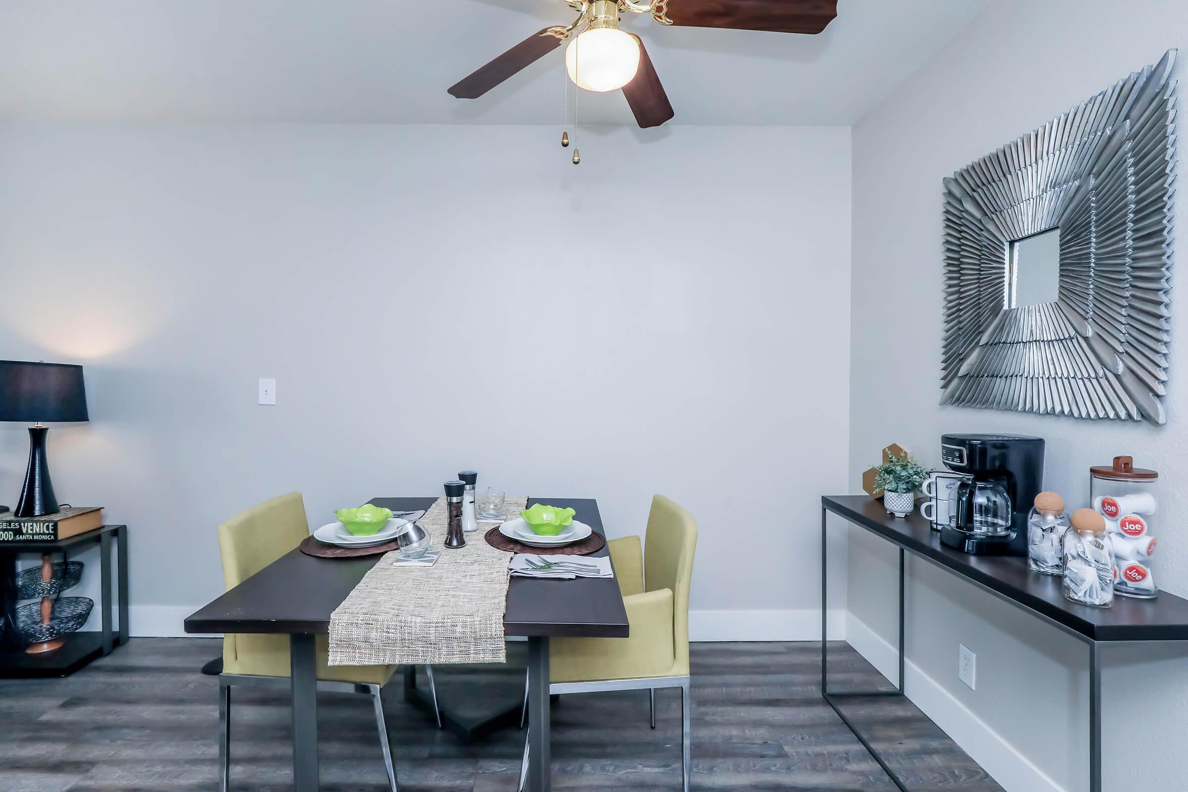 A modern dining area featuring a small rectangular table set with green bowls and napkins. Two yellow chairs are positioned at the table. A decorative mirror hangs on the wall, and a side table with coffee maker and light is visible. A ceiling fan adds a cozy touch to the light-colored room.