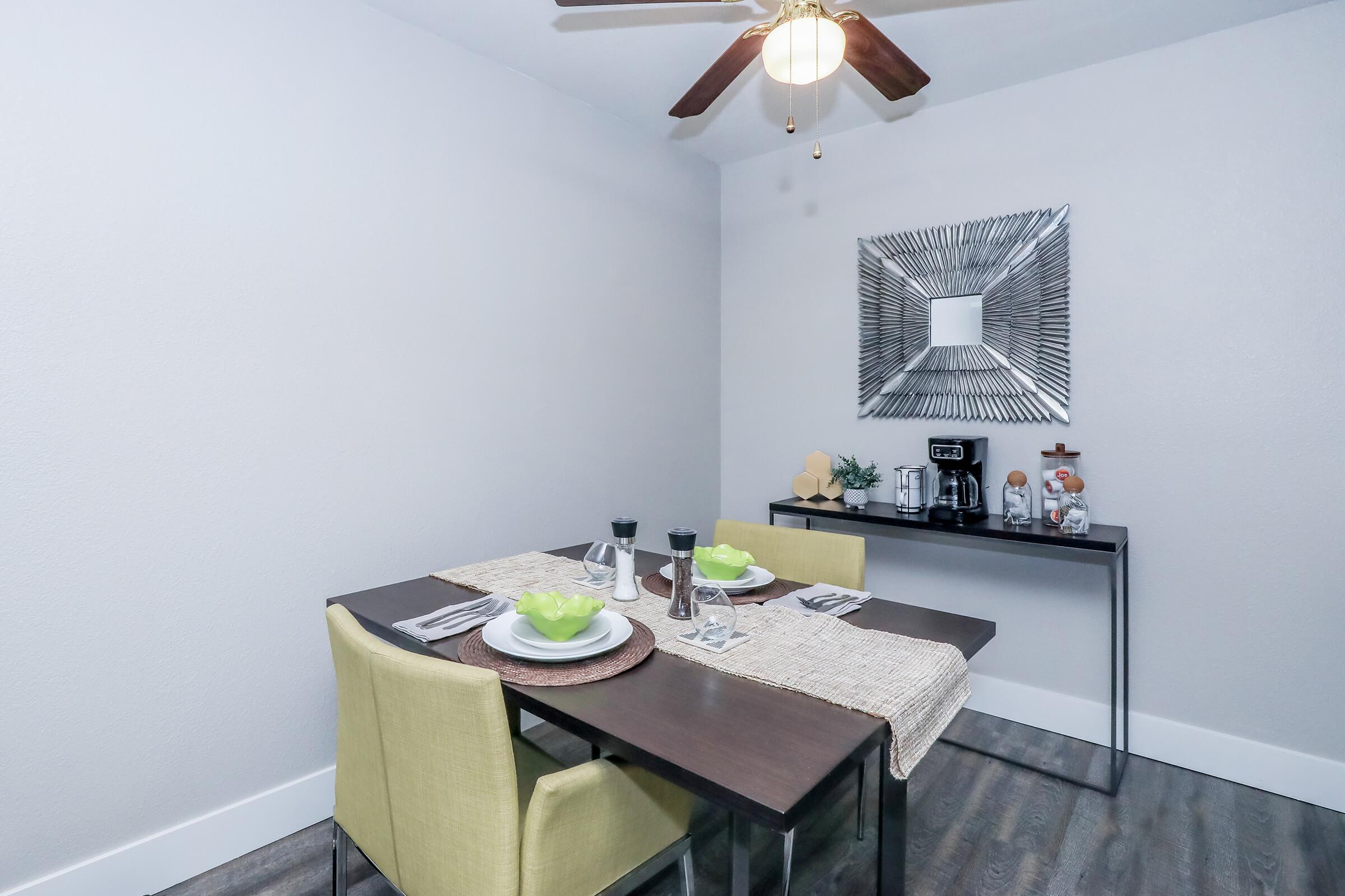 A modern dining area featuring a dark wooden table set for two with plates, utensils, and a glass of wine. Yellow chairs are placed around the table. A woven placemat adds texture, while a decorative mirror hangs on the wall. A coffee maker and decorative items are displayed on a narrow side table.