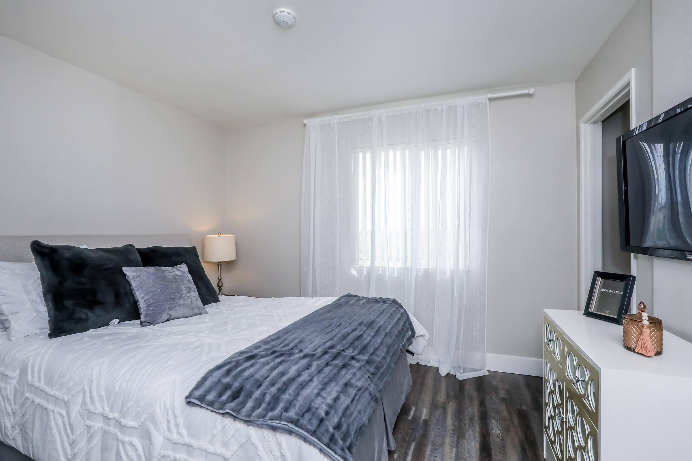 A cozy bedroom featuring a large bed with gray and white bedding, decorative pillows, and a textured throw blanket. A bedside lamp provides soft lighting, while sheer curtains allow natural light to filter in. A wall-mounted TV and a stylish dresser enhance the modern decor.