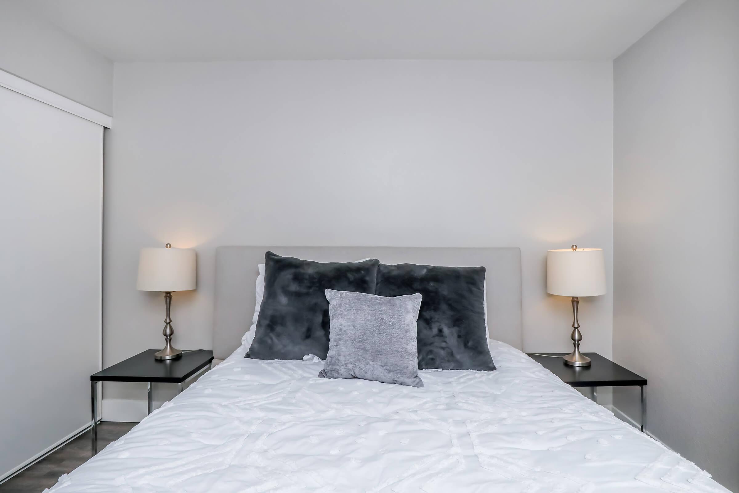 A neatly made bed with a light gray headboard and soft white bedding. A large gray decorative pillow is centered on the bed. On either side of the bed, there are small nightstands with lamps. The walls are painted a light color, creating a calm and minimalist bedroom atmosphere.