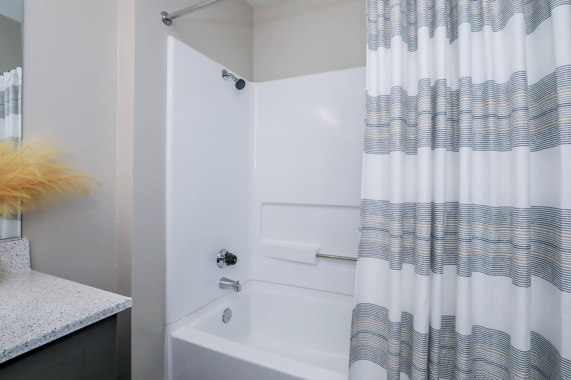 A clean bathroom with a white bathtub and a shower curtain featuring a wavy blue and gray pattern. The toiletries are neatly arranged on the counter, and there is a decorative plant in a vase on the side. The walls are a neutral color, creating a modern and inviting atmosphere.