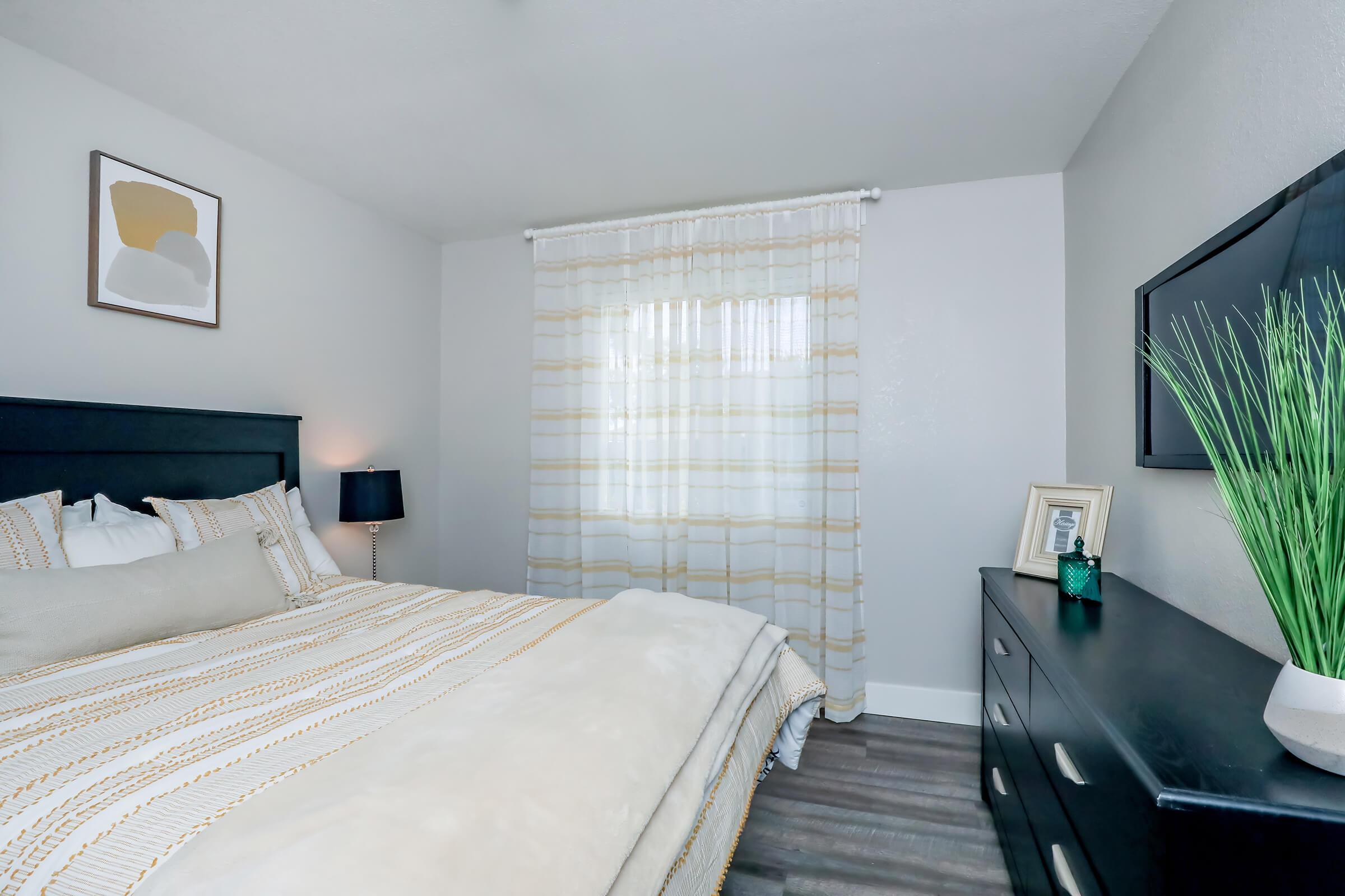 A cozy bedroom featuring a black bed frame with a beige striped comforter, a bedside lamp, and a decorative artwork on the wall. Natural light filters through a sheer striped curtain over a window. A dresser with a plant and photo frame completes the inviting atmosphere.