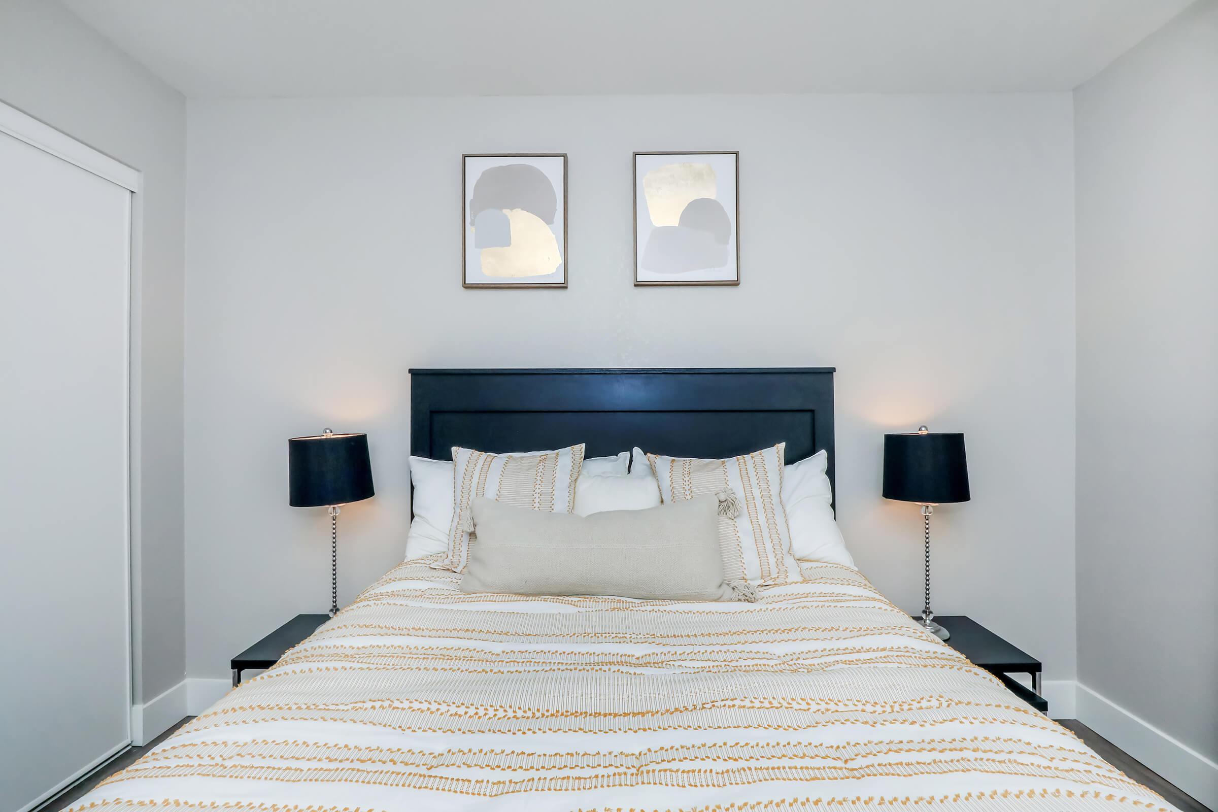 A modern bedroom featuring a neatly made bed with a striped duvet and decorative pillows. Two framed artworks hang above the headboard. Each side of the bed has a black bedside lamp with a white shade, and there is a closet door visible on the left side of the image. The walls are painted light gray.