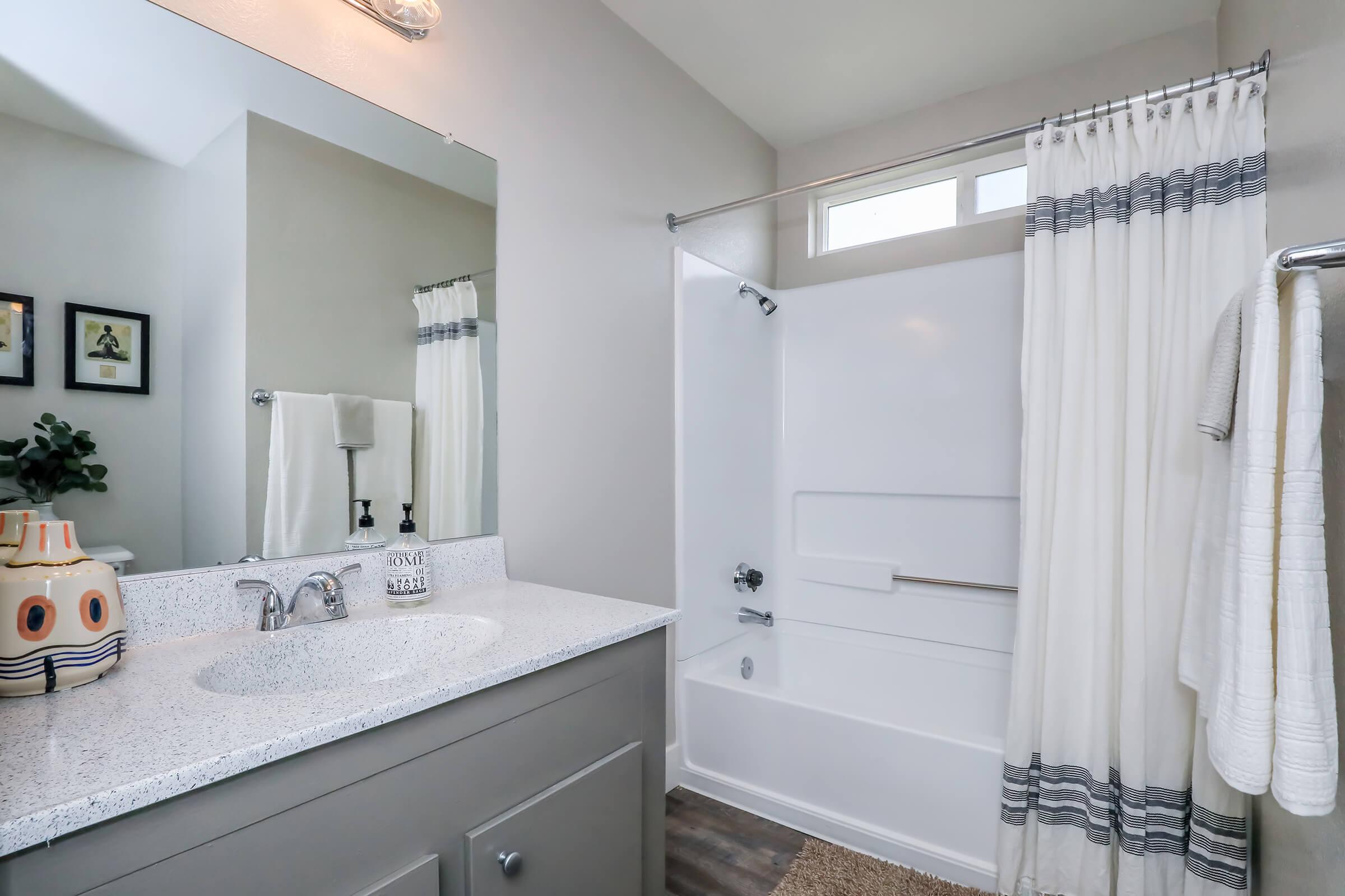 A modern bathroom featuring a white bathtub with a shower, a light gray vanity with a stone countertop, and a large mirrors. Soft white curtains frame the bathtub, and a rug adds warmth to the floor. Decorative elements include a potted plant and framed artwork on the walls.