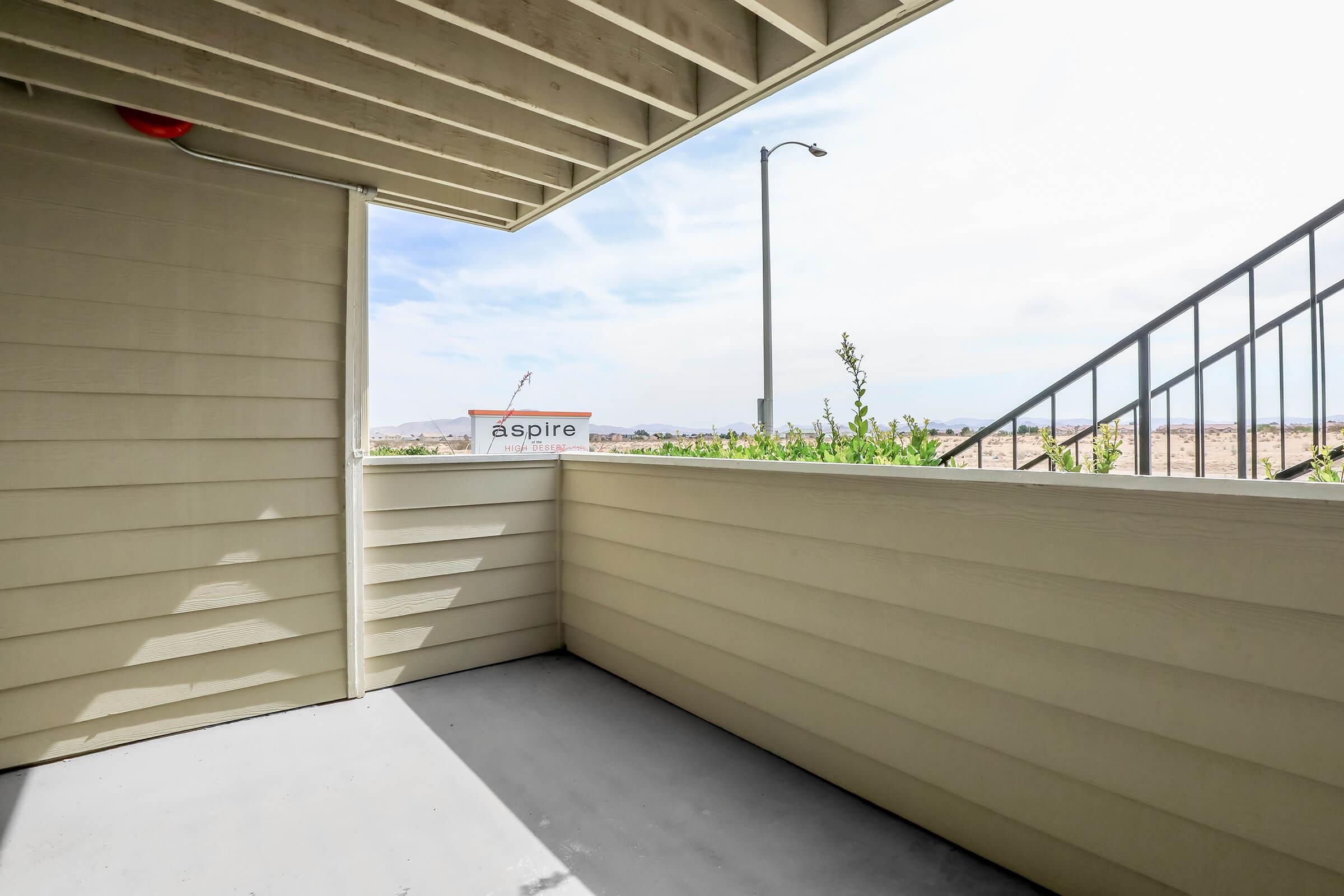 A partially enclosed balcony area featuring beige siding, with a view of the outdoors including a distant landscape, a light pole, and a sign that reads "aspire." The floor is gray and the space is well-lit with natural light coming in.