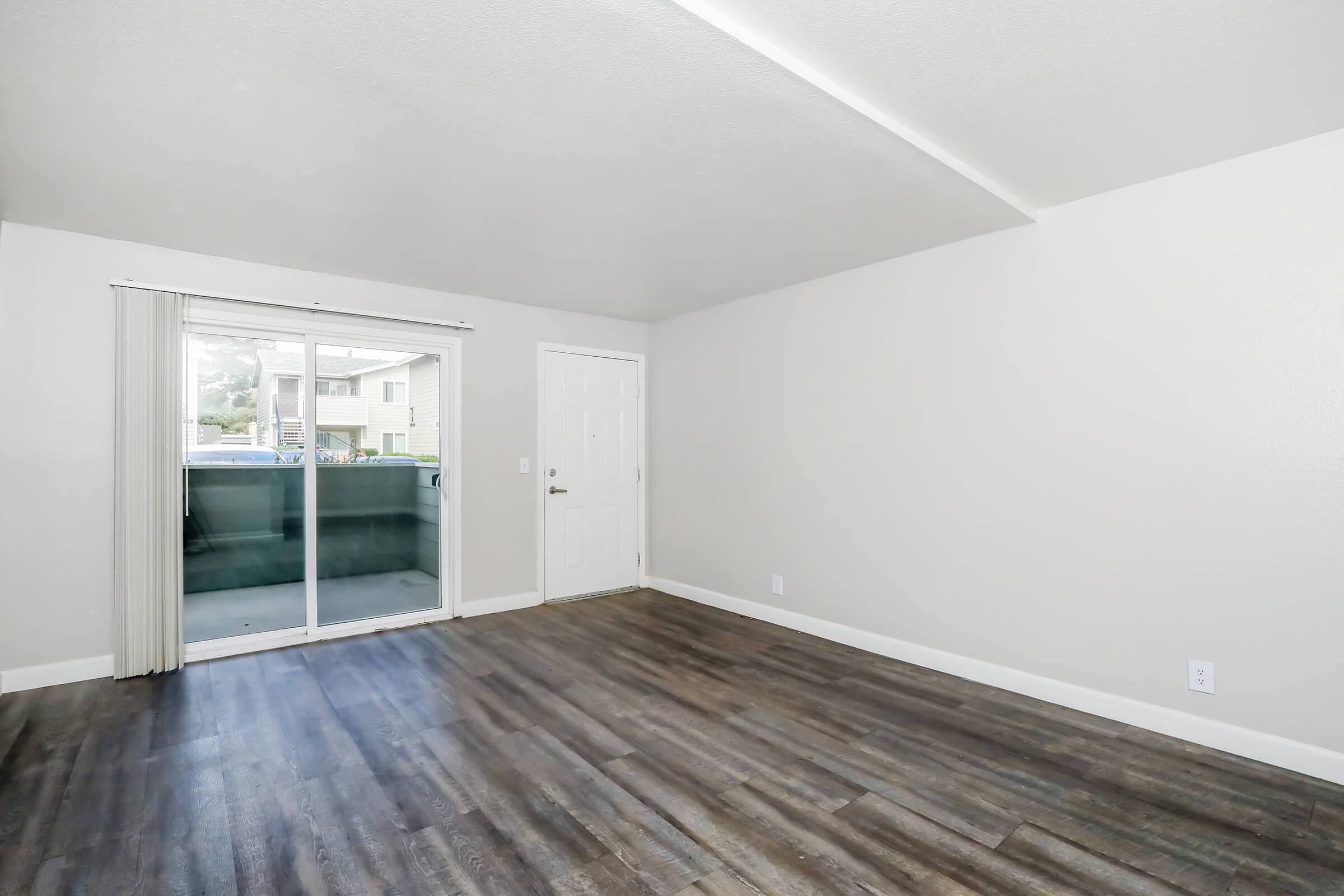 A spacious, empty living room featuring light gray walls and dark wood-like flooring. There is a sliding glass door leading to a balcony, with white curtains on either side. The room has a simple, modern aesthetic with ample natural light.
