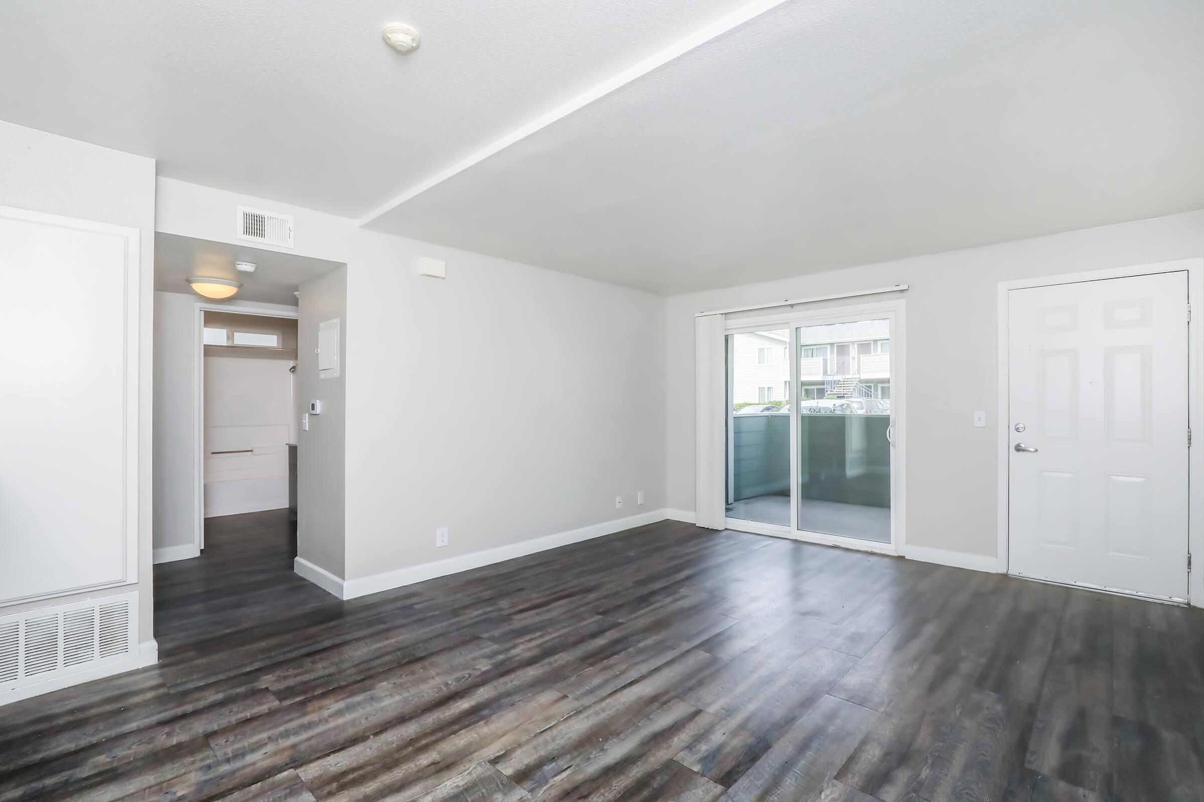 Spacious, empty room featuring light-colored walls and a modern design. The floor is covered with dark laminate, and there is a sliding glass door leading to a balcony. A doorway is visible in the background, leading to another area. Bright, well-lit space ideal for living or leisure.