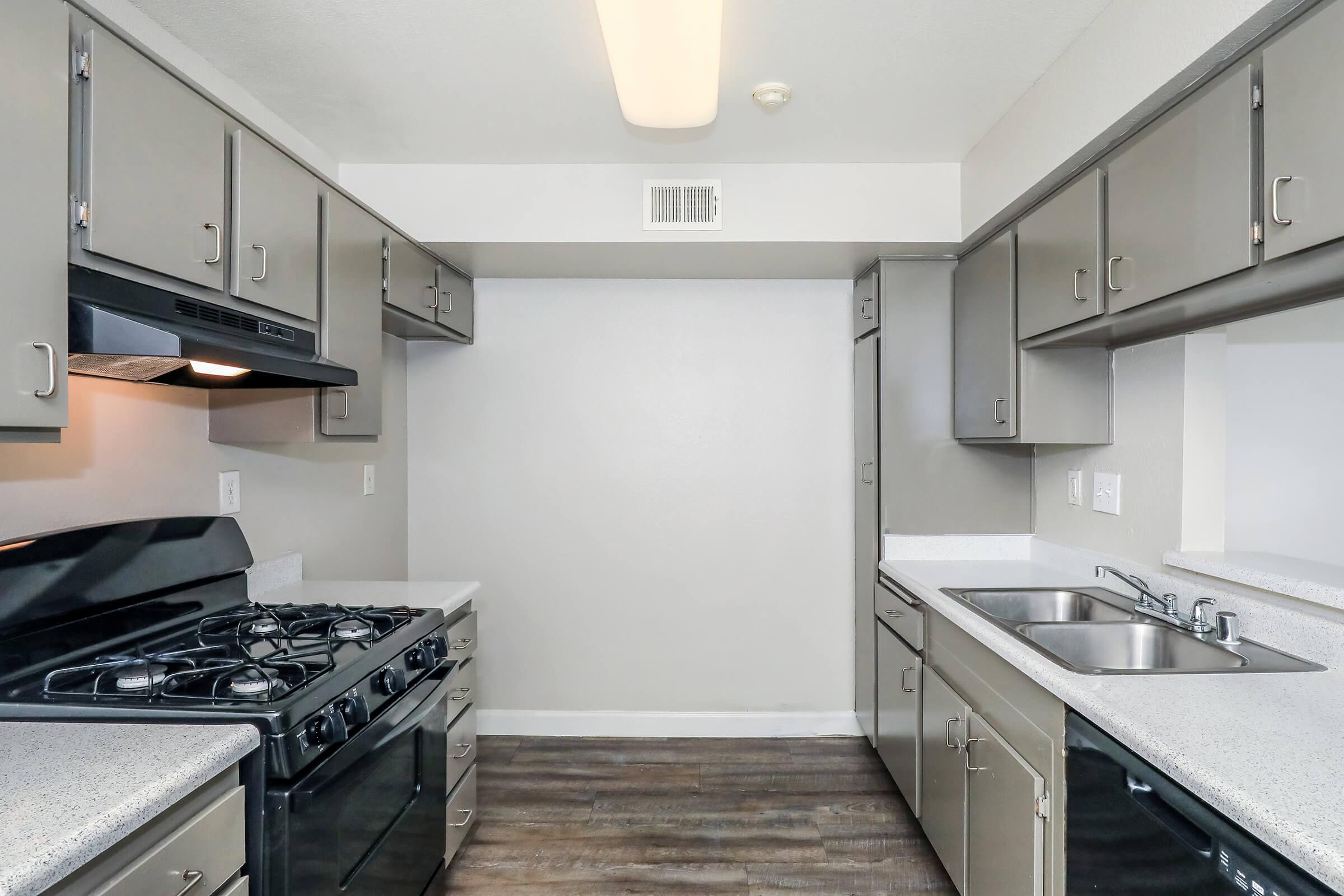 Modern kitchen featuring gray cabinets, a black gas stove, and dual sinks. The space has a neutral wall color and wooden flooring, with ample countertop space and overhead lighting.