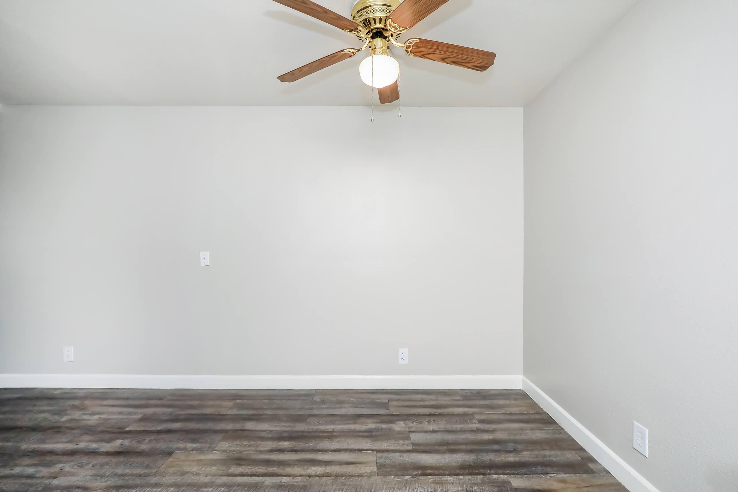 A spacious room with a light-colored wall and wooden flooring. A ceiling fan with wooden blades is centered in the room, providing a bright, airy feel. There are no furniture or decorations present, highlighting the simplicity of the space.