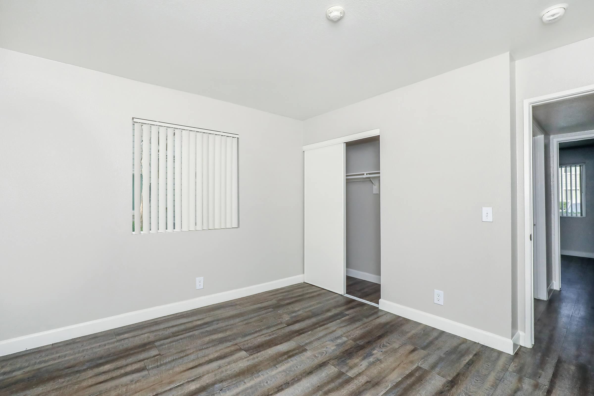 A spacious, empty room featuring light gray walls, a closet with a sliding door, and vertical blinds on a window. The floor is made of dark wood laminate, and there are two doorways leading to adjacent areas. The overall ambiance is bright and minimalist.