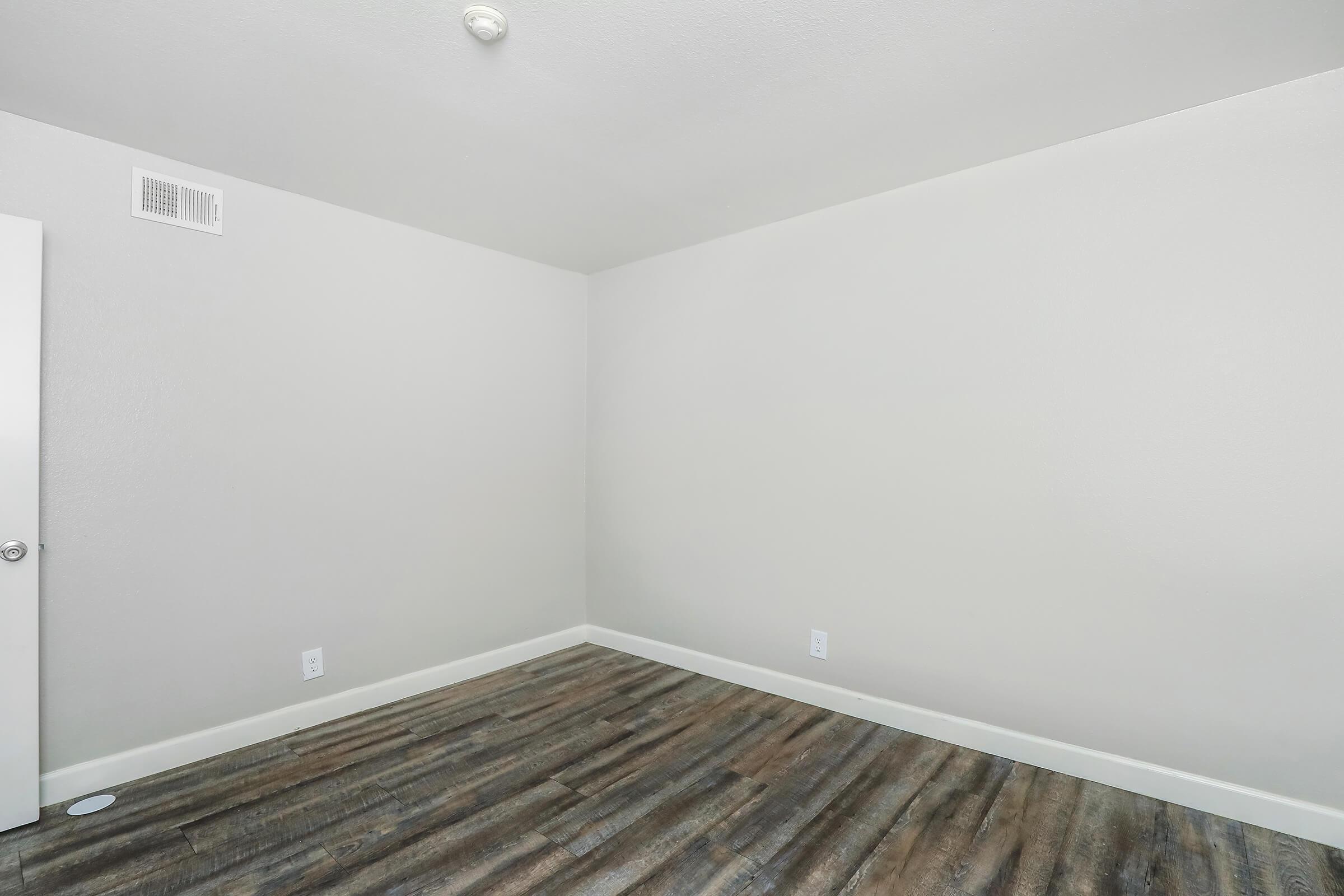 Empty room with light gray walls and a wooden floor. A single white door is visible on the left side of the image, and a ceiling light fixture is present. The space is well-lit and has no furniture or decorations.