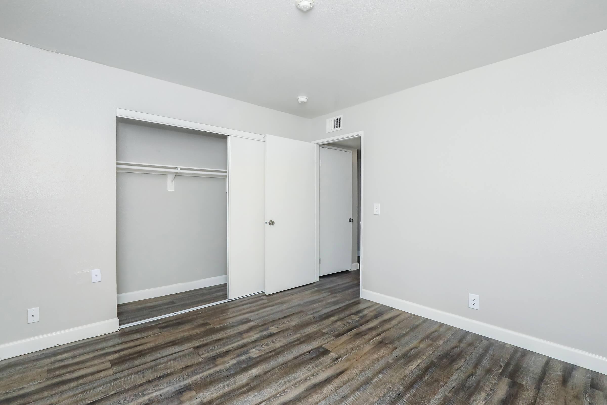 Empty room with light gray walls and wooden flooring. A closet with sliding doors is visible on the left, and there's a door leading to another area on the right. The space is well-lit, featuring a ceiling light and no furnishings.