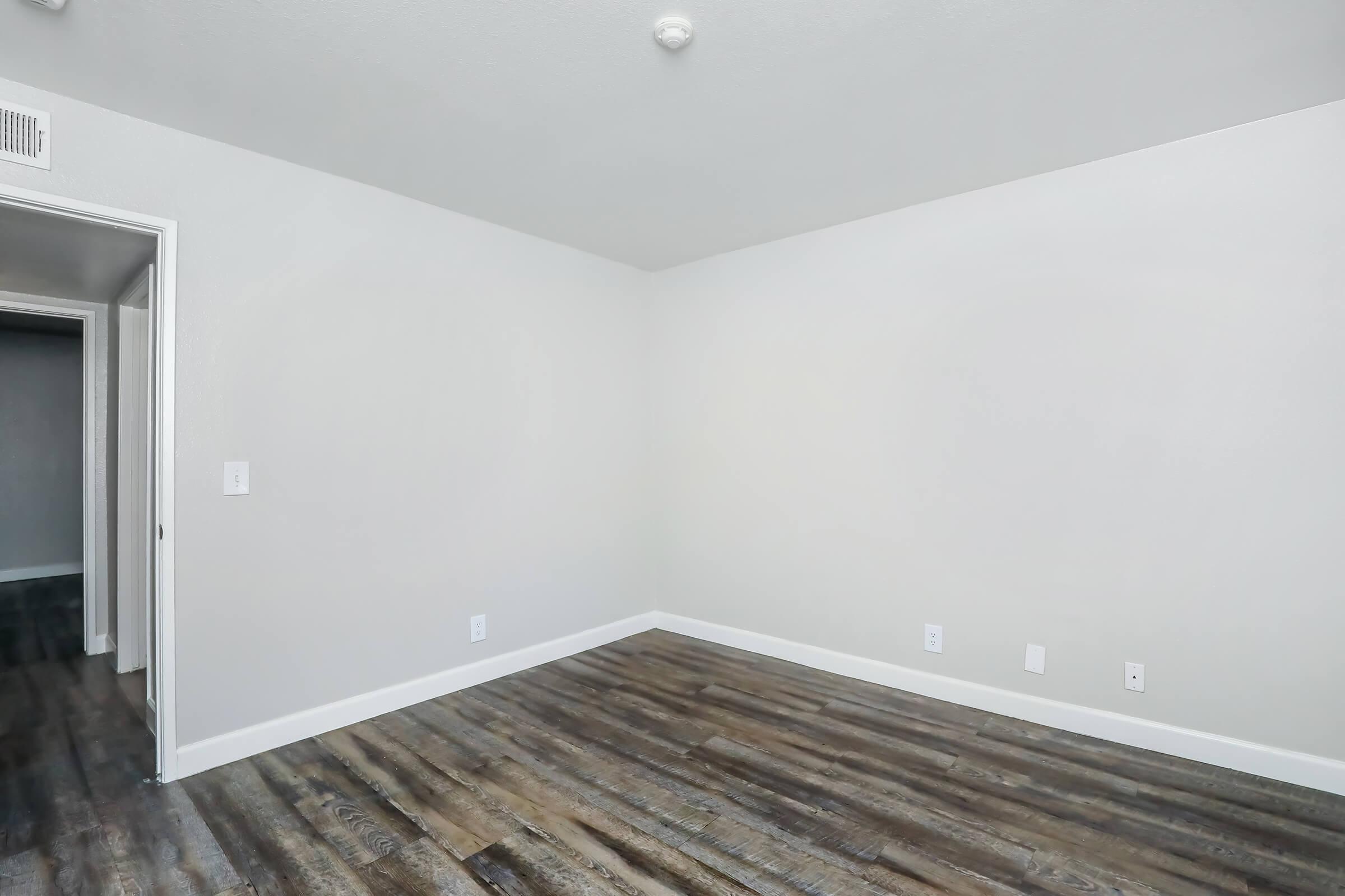 Empty room featuring light gray walls, a smooth ceiling, and dark wood-like laminate flooring. One doorway is visible leading to another room, while the opposite wall is also blank. The space is bright and uncluttered, ideal for various uses or decoration.
