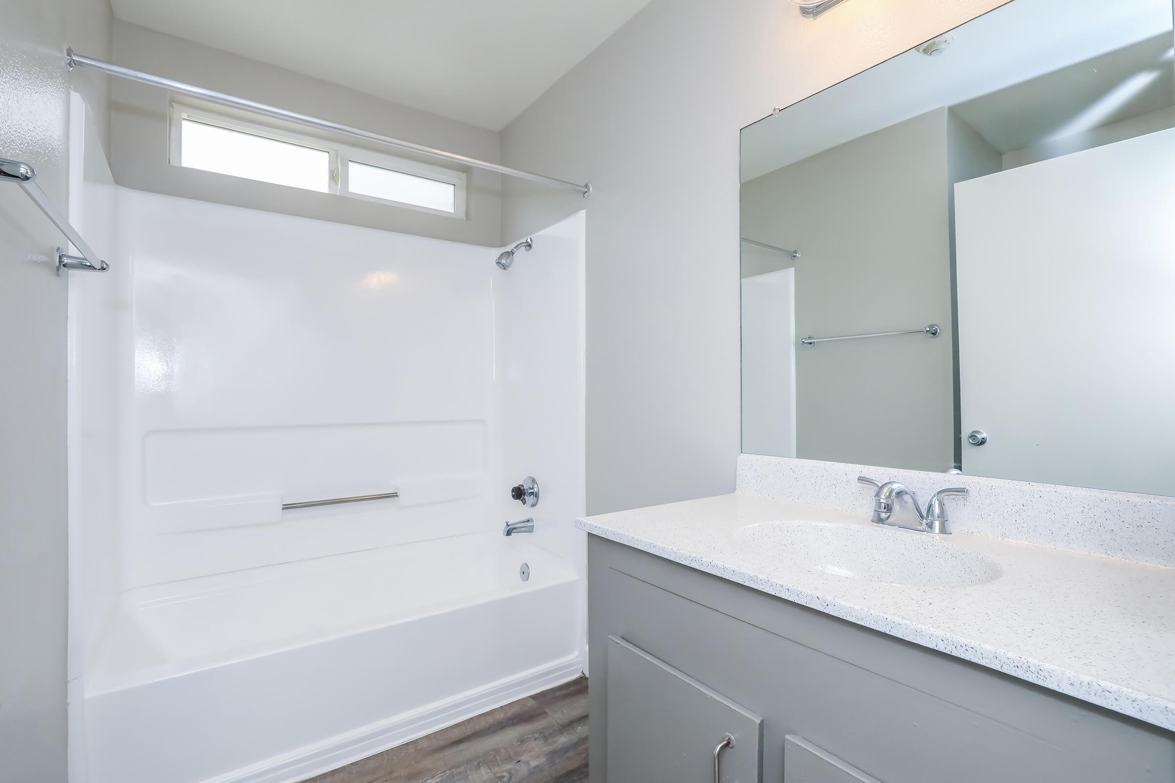 A modern bathroom featuring a white bathtub and shower combination, a light gray vanity with a sink, and a large mirror. Natural light enters through a small window above the bathtub, and the flooring is a neutral wood-like laminate. The walls are painted in a soft, light color.