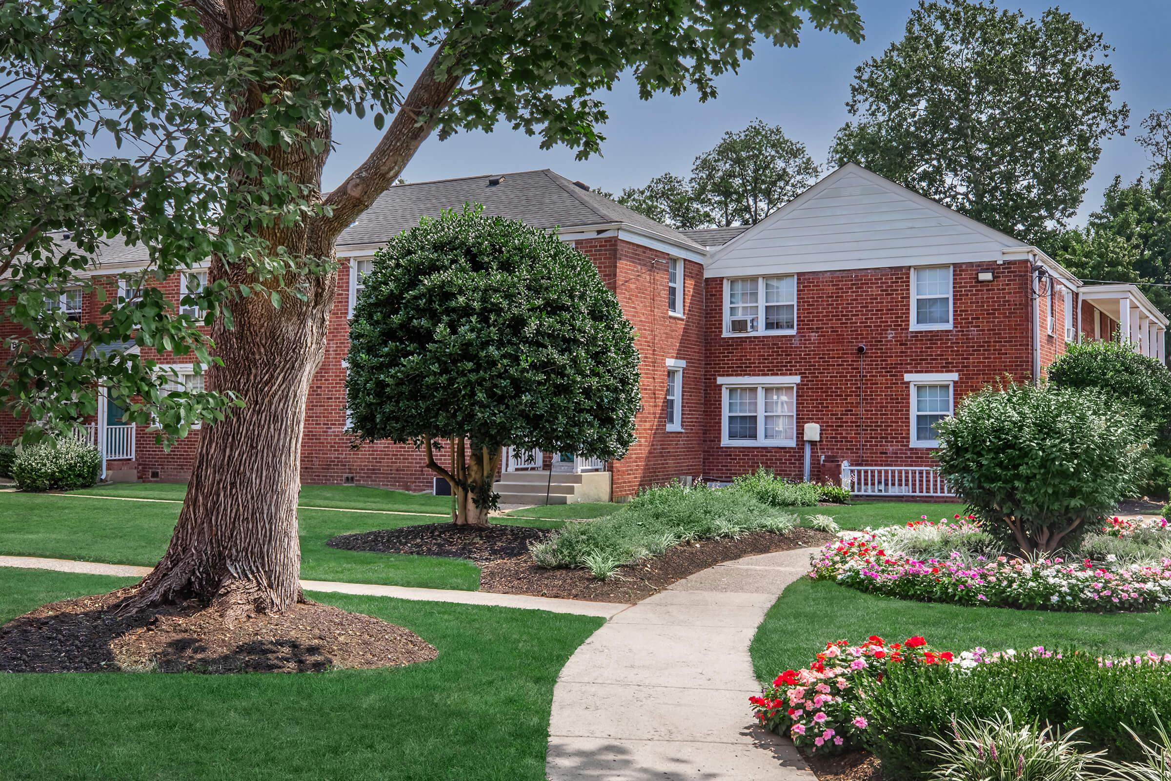 A view of a well-maintained residential area featuring brick apartment buildings surrounded by lush green lawns and colorful flower beds. A large tree stands prominently, with a winding concrete pathway leading through the landscaped grounds.