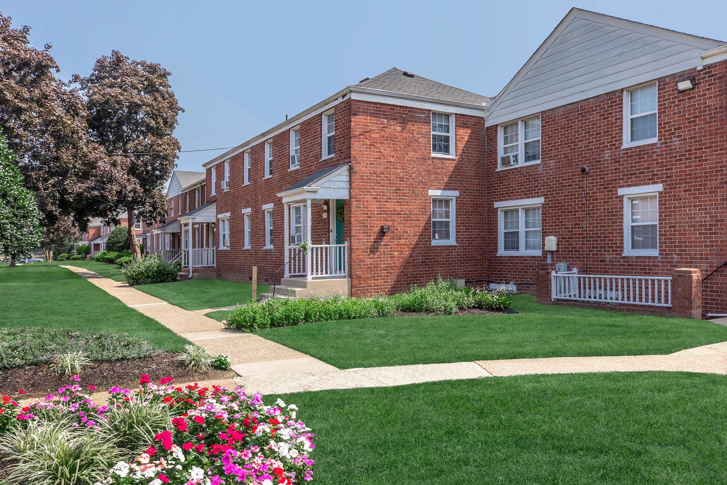 A well-maintained brick apartment building surrounded by green lawns and flower beds. The walkway is lined with neatly trimmed grass and colorful flower arrangements, creating a pleasant outdoor space. Trees can be seen in the background, adding to the neighborhood's charm.