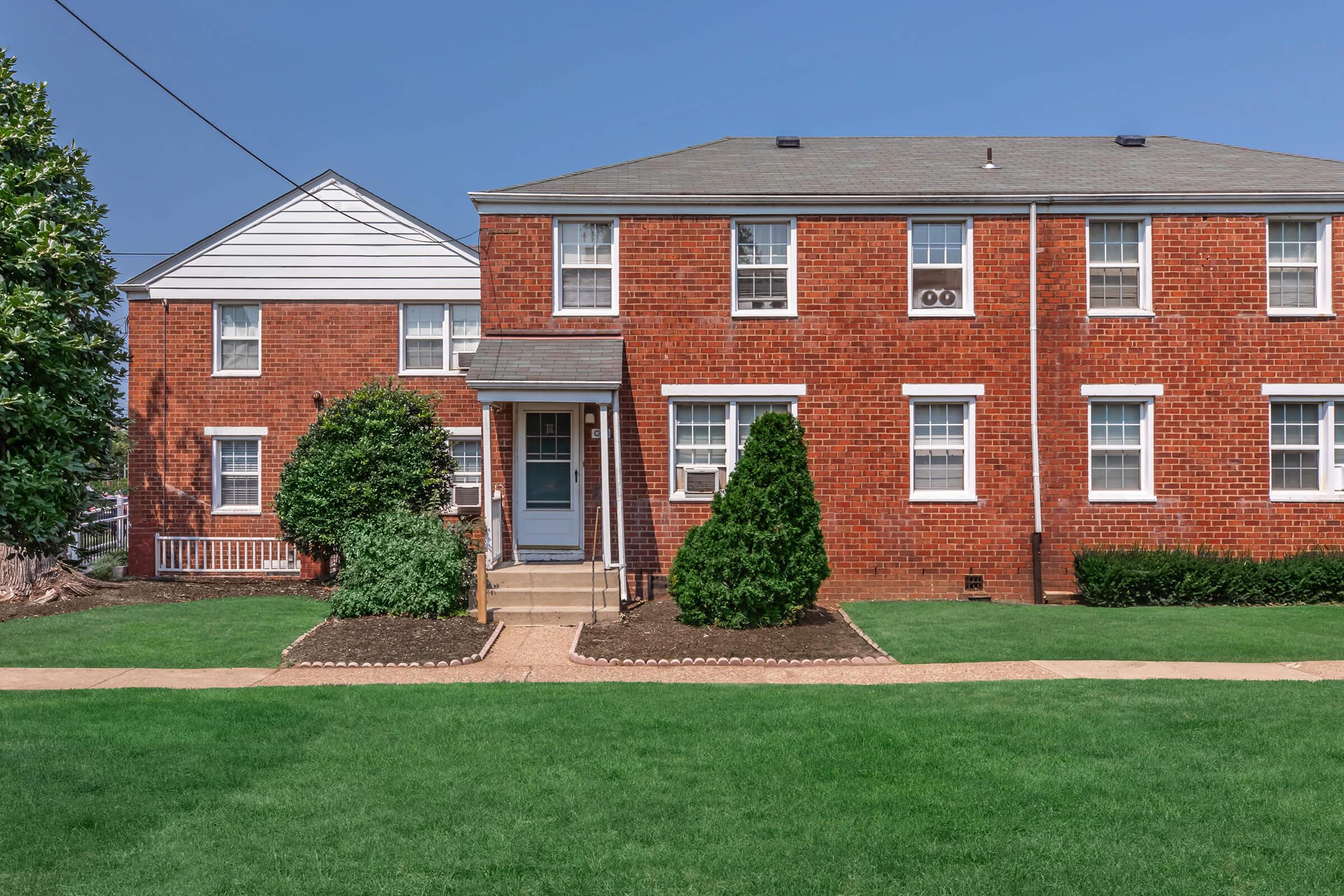 A brick apartment building with a well-maintained lawn. The structure features two sections: a two-story red brick building with white-framed windows and a smaller, white-paneled section. A concrete path leads to the front door, and there are neatly trimmed bushes on either side of the entrance.