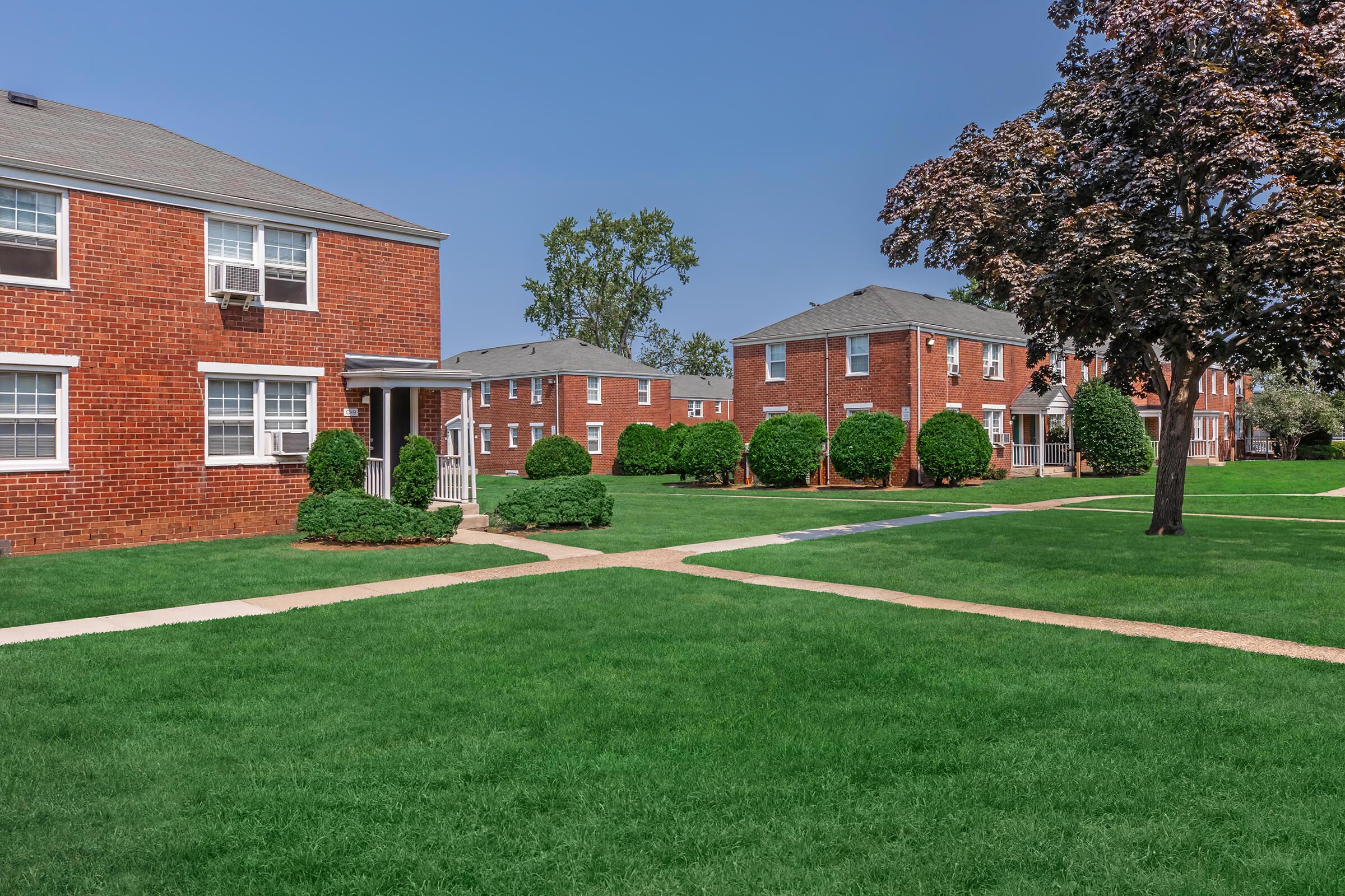 A well-maintained apartment complex featuring red-brick buildings surrounded by lush green lawns and neatly trimmed bushes. Pathways lead through the yard, creating a welcoming atmosphere under a clear blue sky.