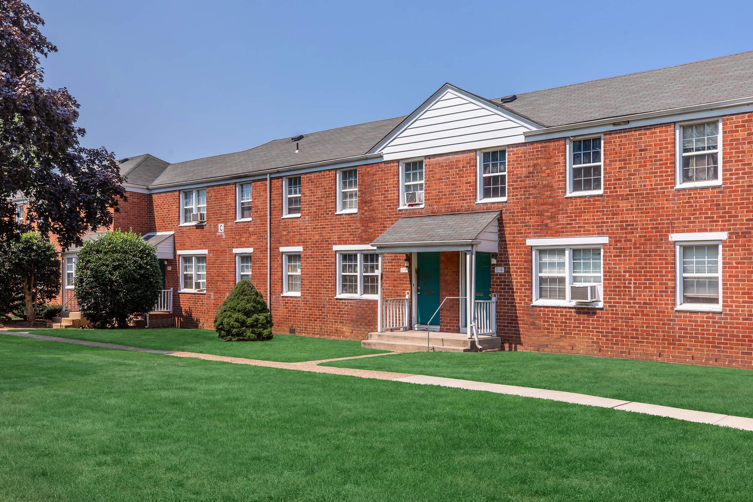 A row of brick apartment buildings with white trim, featuring multiple windows and front entrances. The well-maintained green lawn is visible in front of the buildings, and there are small trees and bushes on either side of the pathway leading to the doors. The sky is clear and blue.