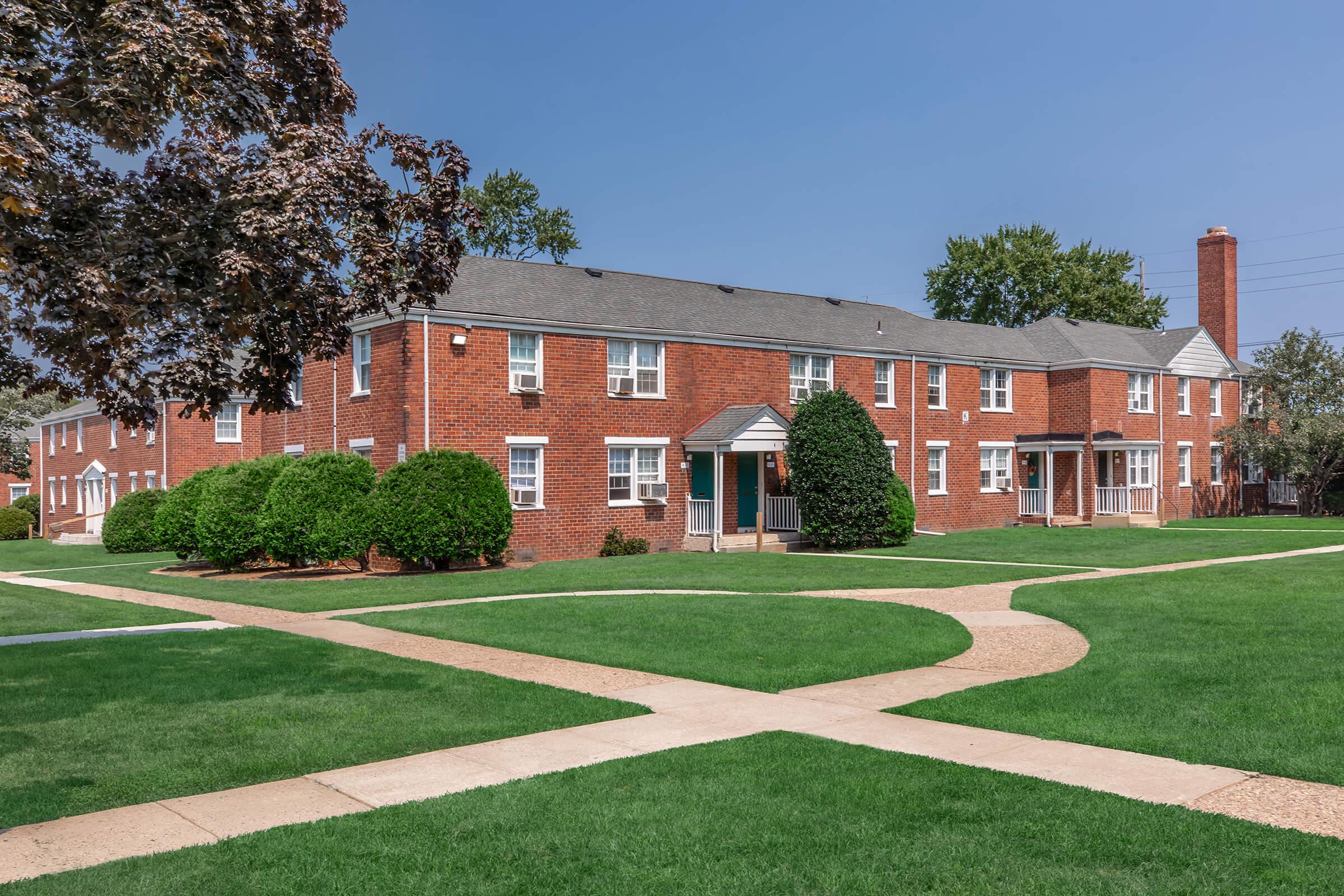 A well-maintained brick apartment complex surrounded by green lawns and neatly trimmed bushes. Sidewalks create intersecting pathways leading to the building entrances under a clear blue sky.