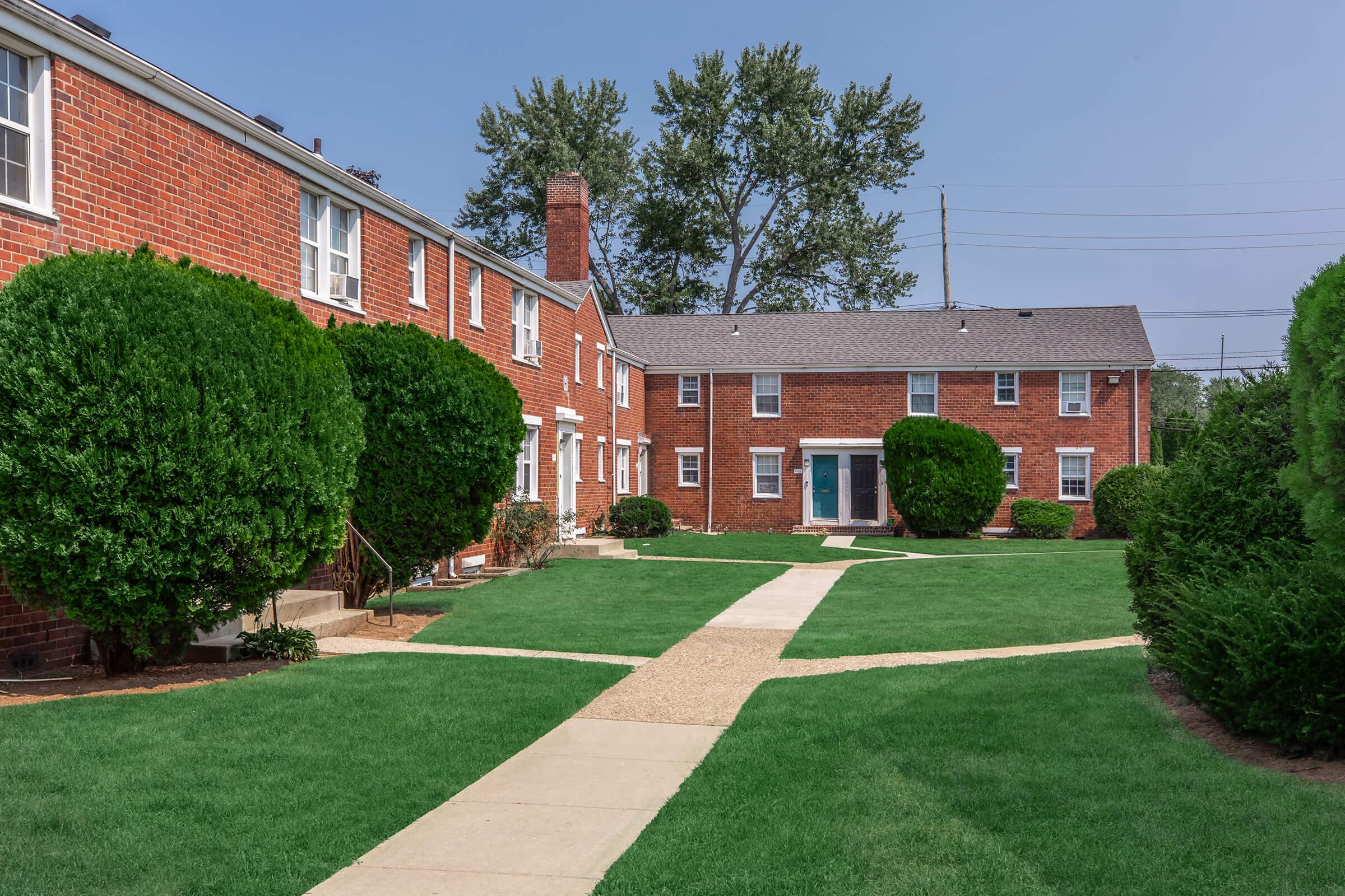 Brick buildings with landscaped greenery and manicured lawns. A wide path winds through the grass, leading between the buildings. The scene features neatly trimmed bushes and trees in the background, under a clear blue sky.