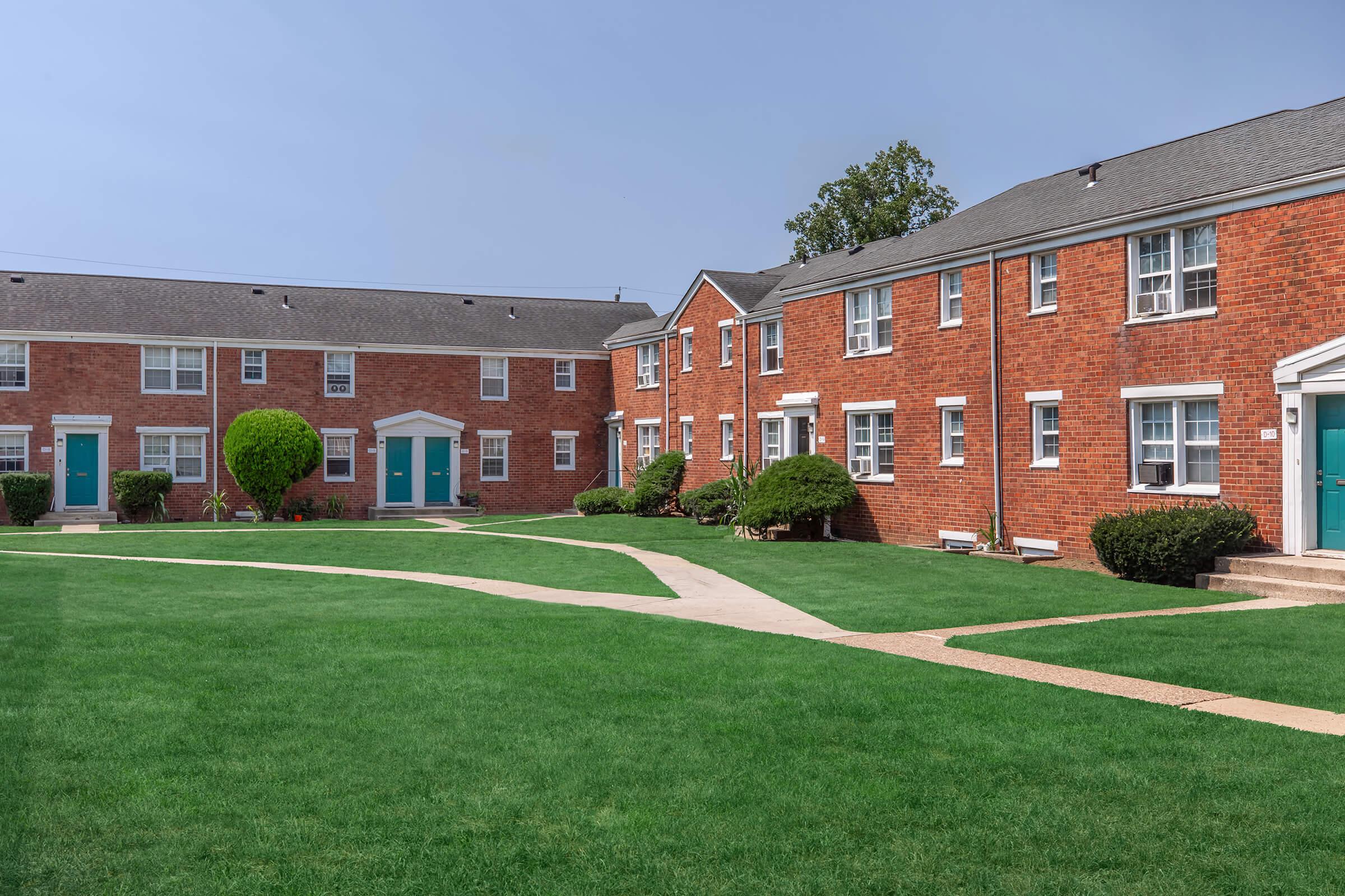 A sunny view of a residential complex featuring brick buildings with teal doors, surrounded by well-maintained lawns and shrubs. Sidewalks curve through the grassy area, leading to the entrances of the apartments. The atmosphere is peaceful and inviting, typical of suburban living.