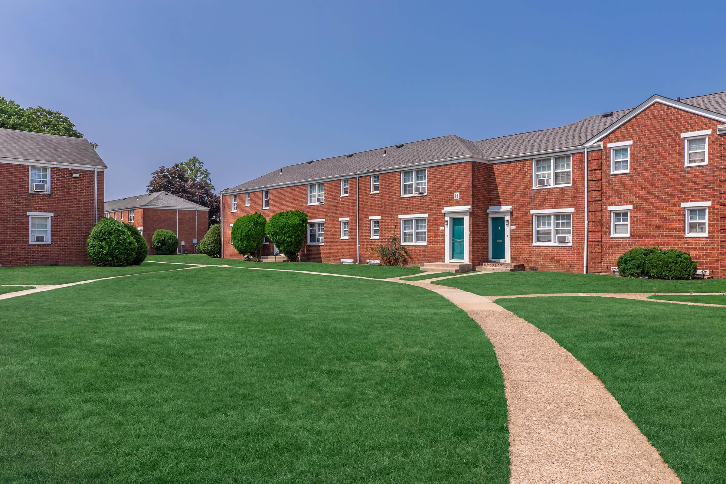 A well-maintained residential complex featuring red brick buildings arranged around a grassy lawn. The pathway curves through the green space, connecting the units, with decorative shrubs adding to the landscape. Clear blue sky overhead creates a bright and inviting atmosphere.