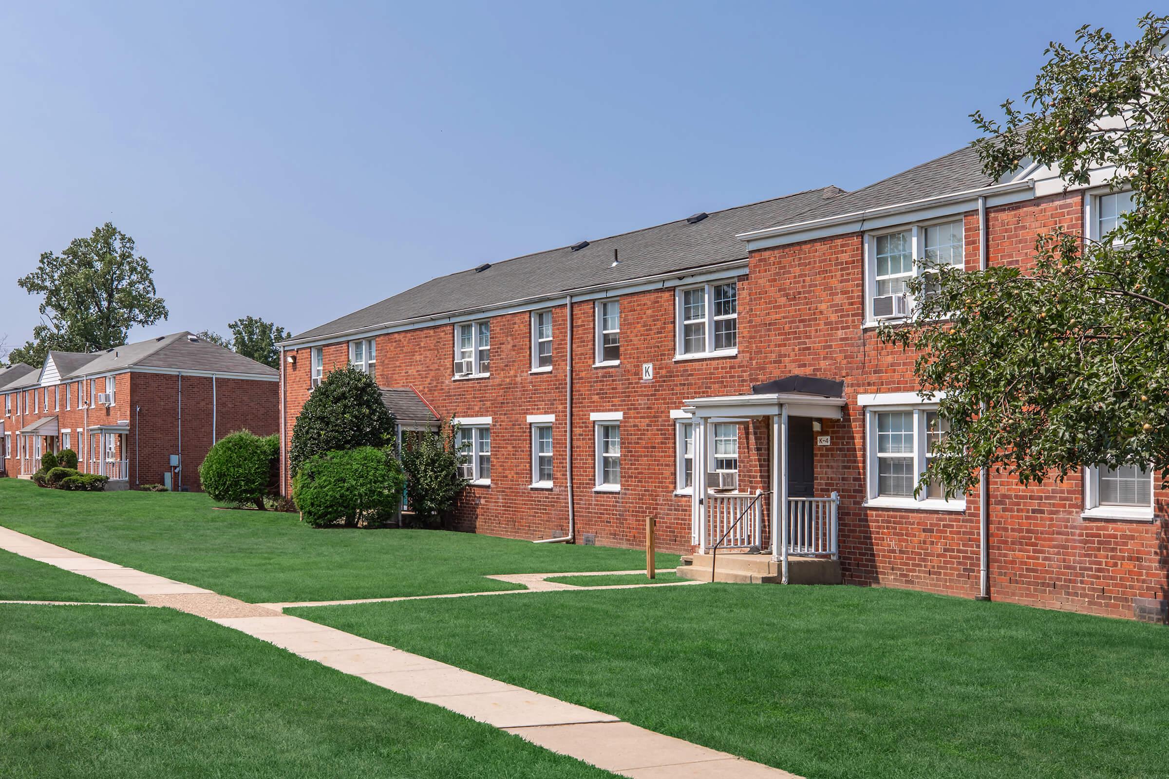 A row of red brick apartments with multiple units, surrounded by well-maintained green lawns. The pathway leads to the front entrance of one building, while trees and shrubs add to the landscaped area. The sky is clear and blue, indicating a sunny day.