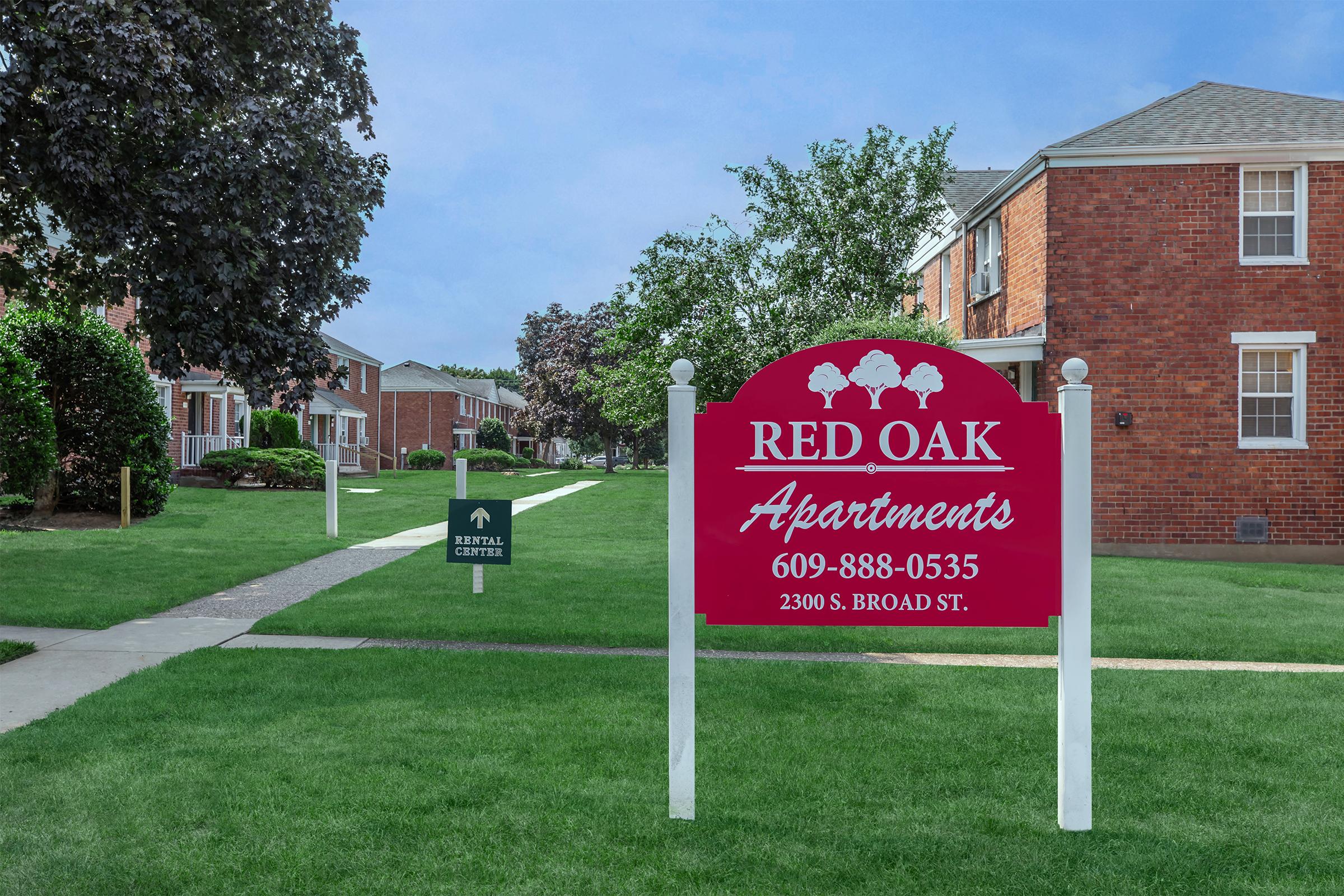 A vibrant red sign with white text reading "RED OAK Apartments" and a phone number, positioned in a green lawn area in front of brick apartment buildings. The background features trees and a clear blue sky. A smaller sign to the left indicates a rental office.