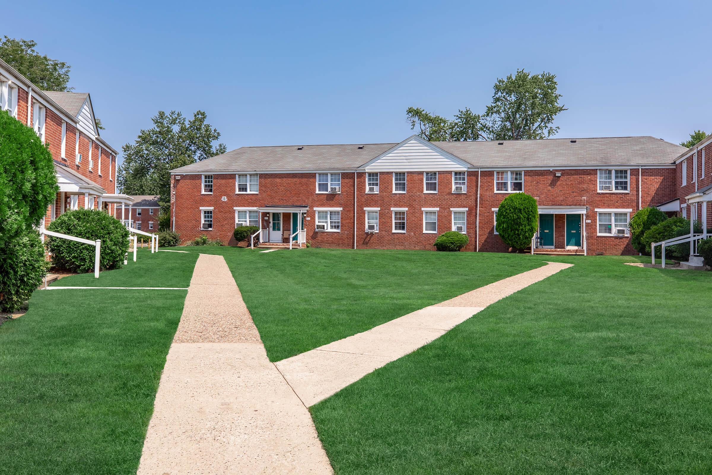 A view of a residential area featuring brick apartment buildings with multiple units, set around a well-maintained green lawn. Pathways made of concrete intersect the grassy area, lined with shrubs and small trees, under a clear blue sky. The scene conveys a peaceful, suburban environment.